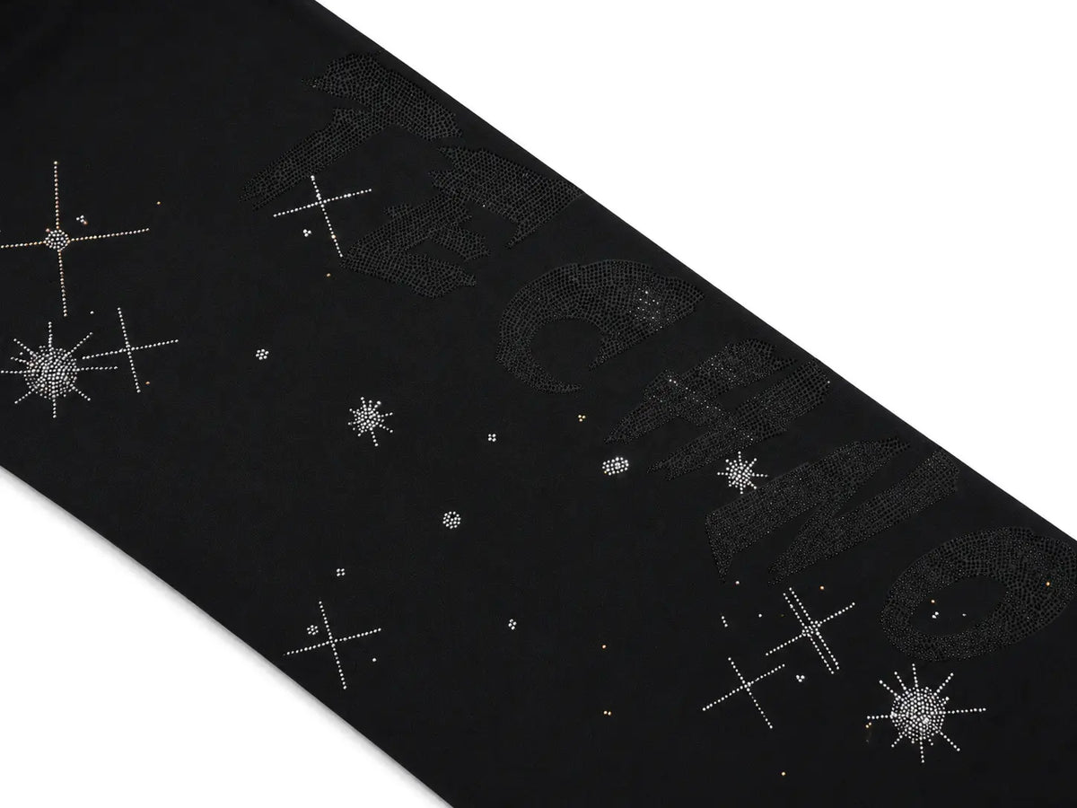 A close-up of a black fabric accessory, likely a belt or scarf, featuring silver rhinestone starburst and cross motifs with a subtle tonal brand logo in the background. TCH Celestial Rhinestone Wide-Leg Sweatpants