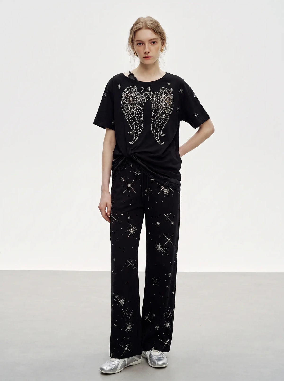 A model stands in a studio wearing a black short-sleeve T-shirt with a large silver wing graphic and scattered star-like sparkles, paired with matching black wide-leg pants featuring star prints, and silver sneakers. TCH Celestial Rhinestone Wide-Leg Sweatpants