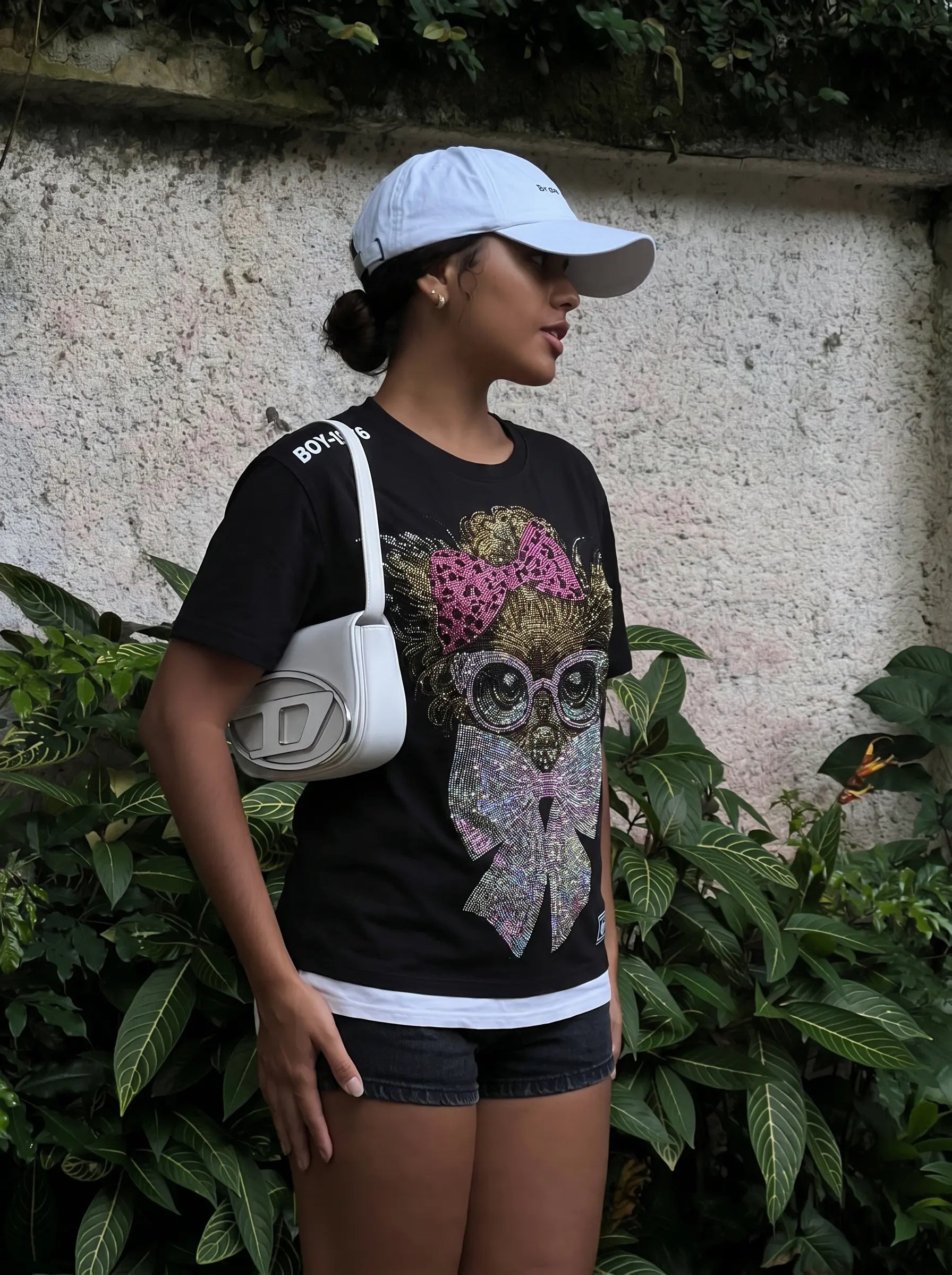 Person wearing a black t-shirt with a colorful graphic design, white cap, and white bag, standing against a wall with greenery. TCH Chihuahua Bow Rhinestone T-Shirt