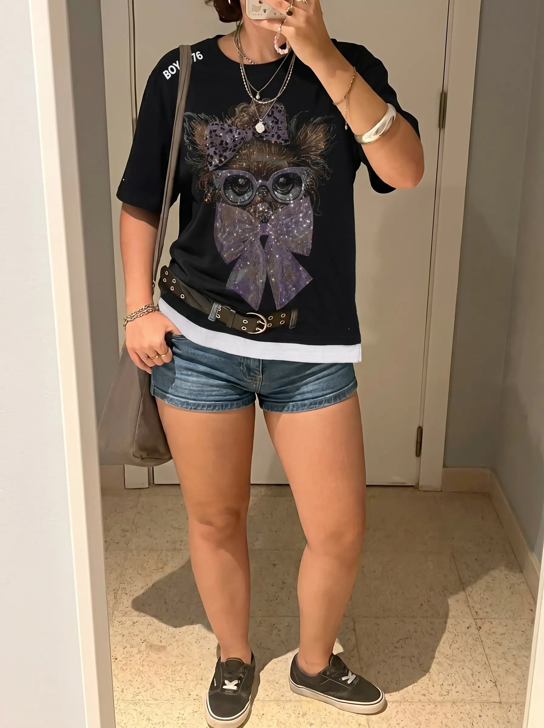 Person wearing a black t-shirt with a graphic design, denim shorts, and black sneakers in a room. TCH Chihuahua Bow Rhinestone T-Shirt