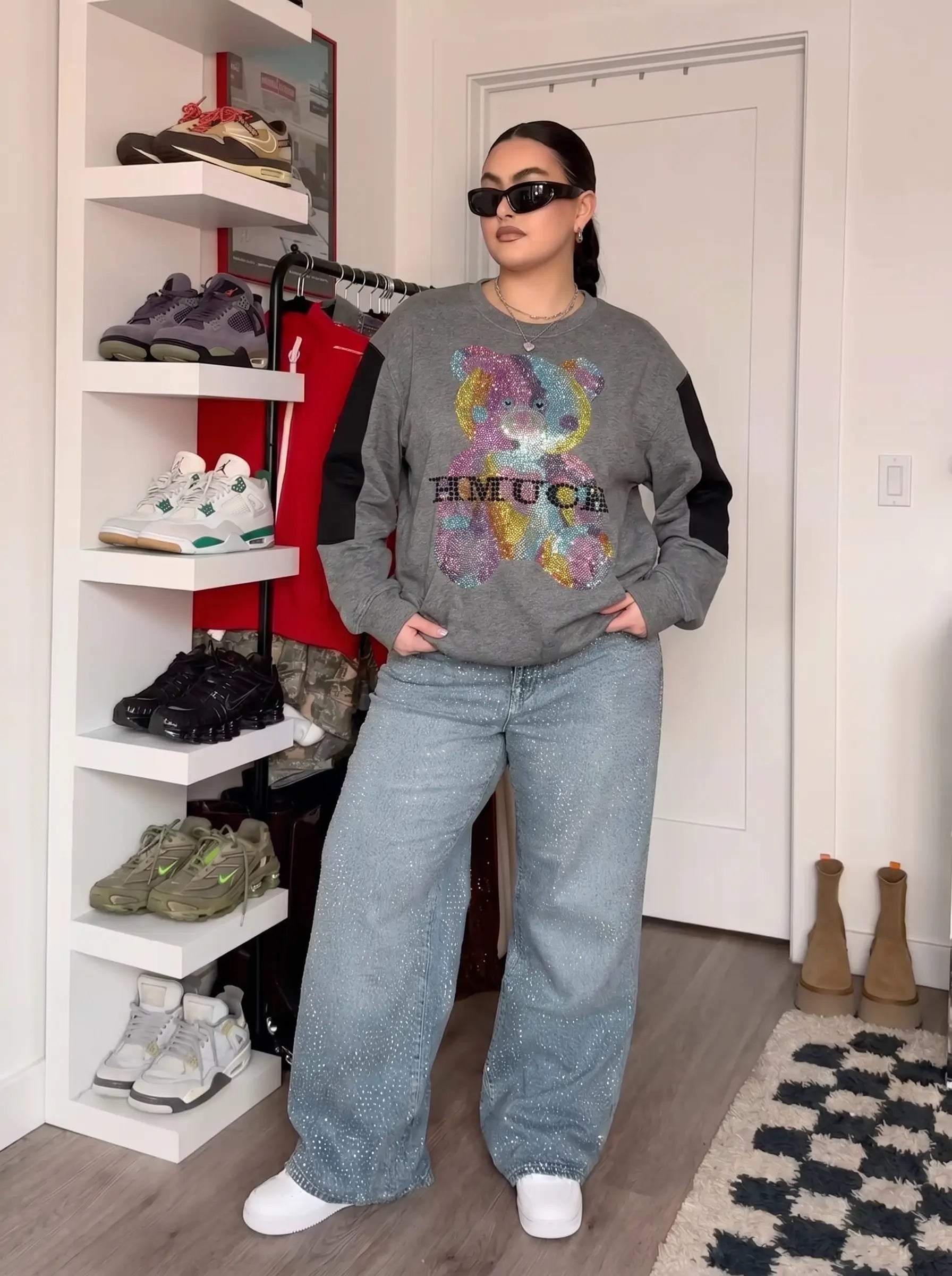A woman modeling a gray sweatshirt with colorful graphic print and black sleeves, paired with light blue wide-leg jeans and white sneakers, standing in a room with a shoe rack and door in the background. TCH Colorful Rhinestone Teddy Bear Crewneck Sweatshirt