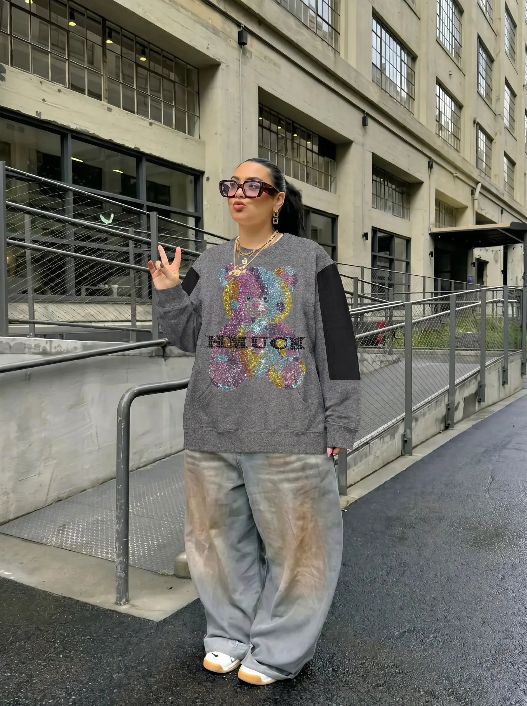 A person standing outdoors in front of a concrete industrial building, wearing a gray sweatshirt with a colorful graphic and the word 'EMUCH' across the chest, paired with loose gray pants and white sneakers. TCH Colorful Rhinestone Teddy Bear Crewneck Sweatshirt