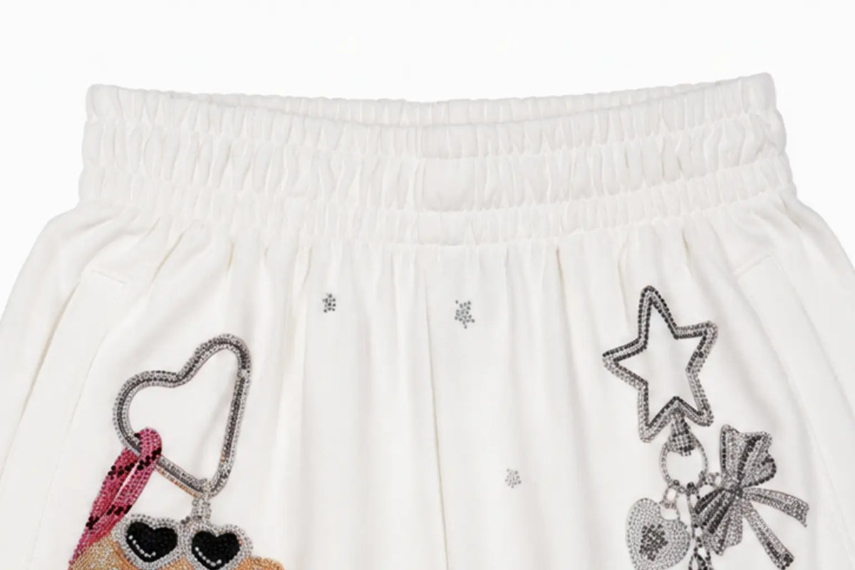 Close-up of a white or off-white fabric garment, likely a skirt or shorts, featuring an elastic waistband and decorative embellishments including a silver beaded star, a pink and black heart charm, and small silver star details. TCH Crystal Bear Graphic Wide-Leg Sweatpants