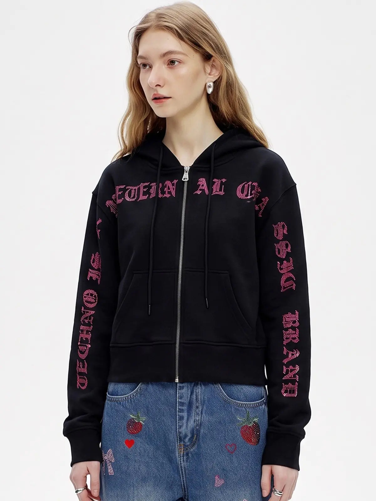 A woman wearing a black zip-up hoodie with pink gothic text graphics on the chest and sleeves, paired with blue jeans featuring strawberry and heart patches, against a plain white background. TCH Crystal Eagle Graphic Zip Up Hoodie