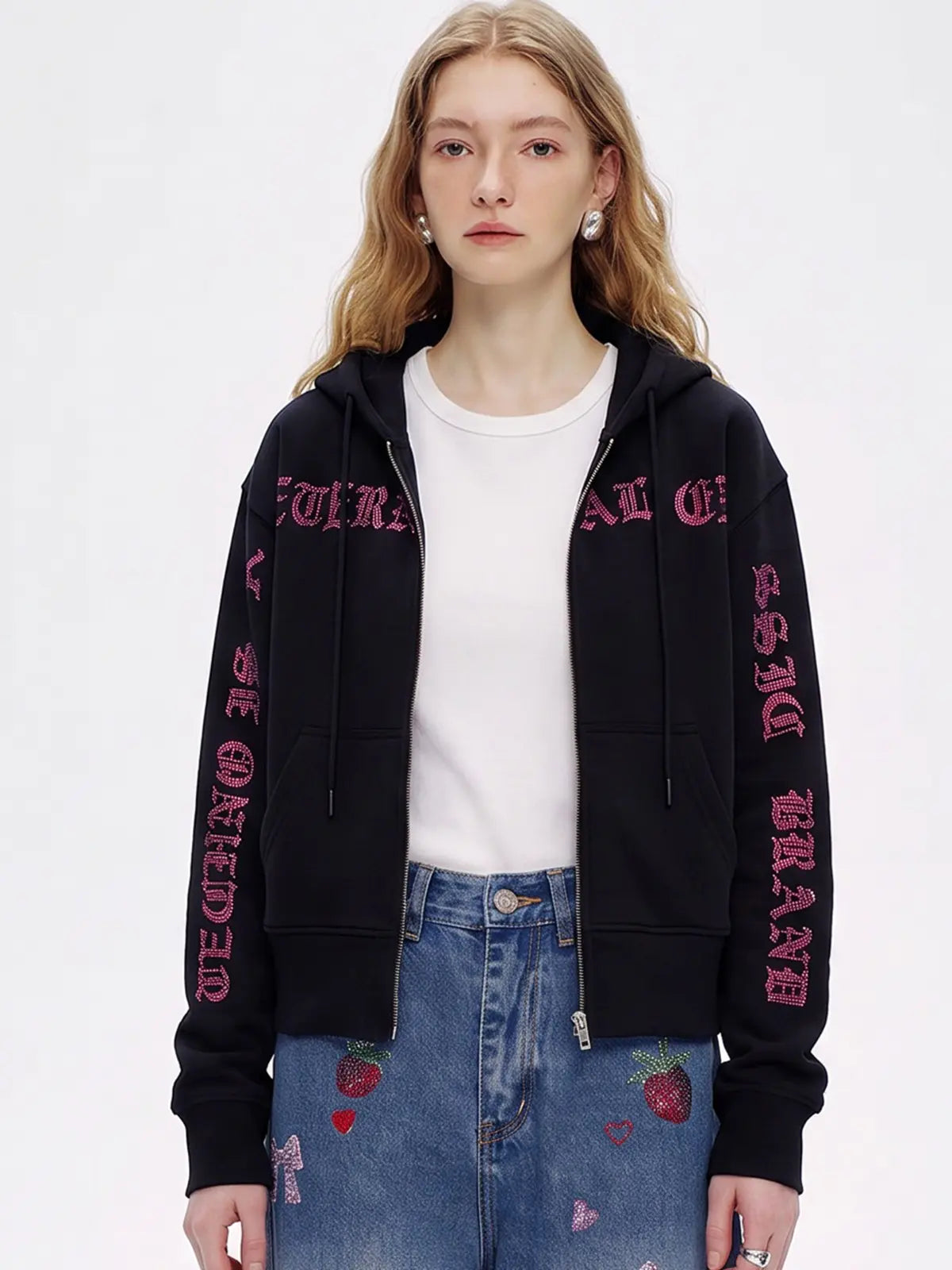 A woman modeling a black zip-up hoodie with pink gothic-style text printed down both sleeves, worn over a white t-shirt and blue jeans with strawberry patches. TCH Crystal Eagle Graphic Zip Up Hoodie