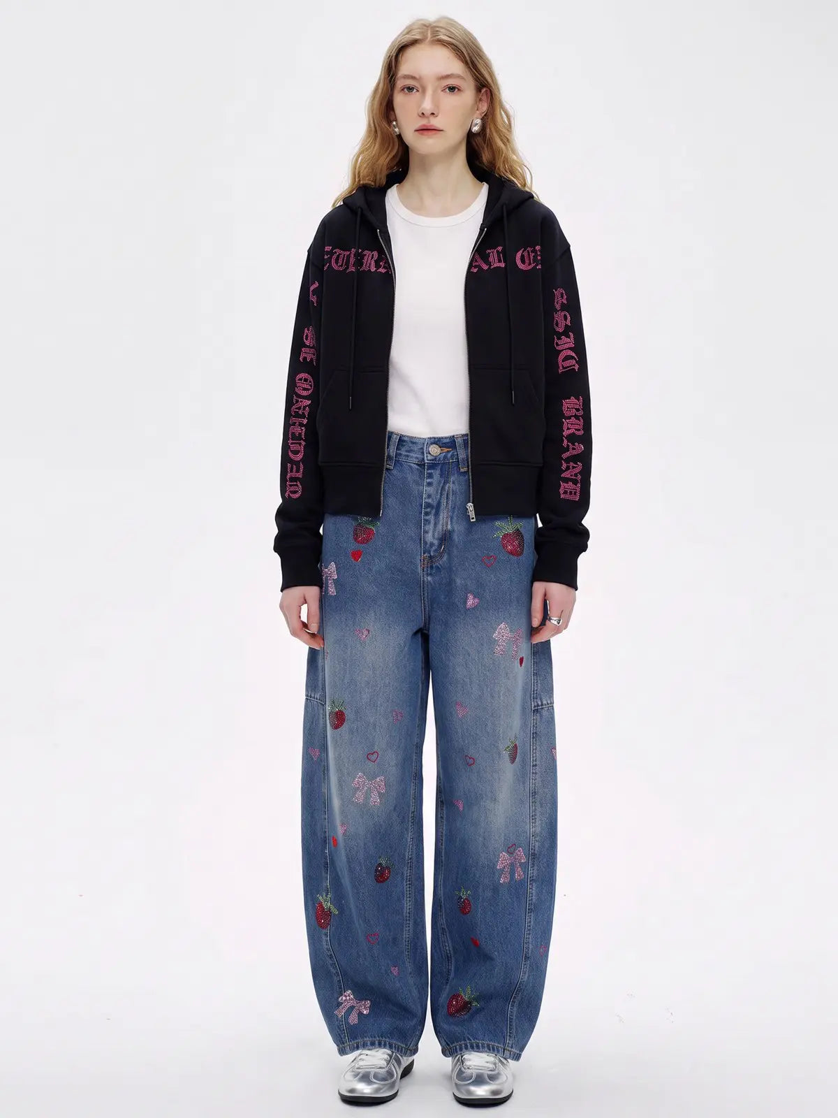 A model stands against a white studio background wearing a black zip-up hoodie with pink gothic text down the sleeves, a white crewneck top, wide-leg blue jeans with pink bow and strawberry prints, and silver sneakers. TCH Crystal Eagle Graphic Zip Up Hoodie