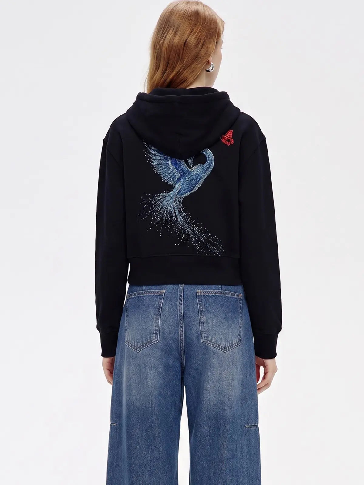 A model viewed from the back wearing a black cropped hoodie with a large blue bird graphic and a small red rose detail on the back, paired with wide-leg blue jeans against a plain white background. TCH Crystal Eagle Graphic Zip Up Hoodie