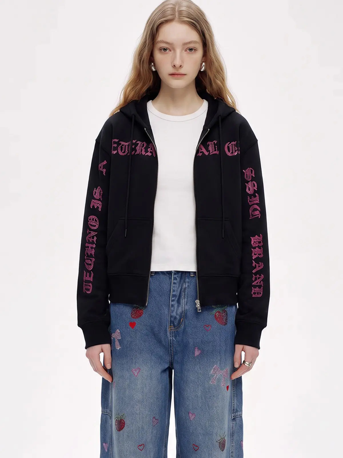 A model wearing a black zip-up hoodie with pink gothic text embroidery on the sleeves, styled over a white t-shirt and blue jeans with strawberry prints, against a plain white background. TCH Crystal Eagle Graphic Zip Up Hoodie