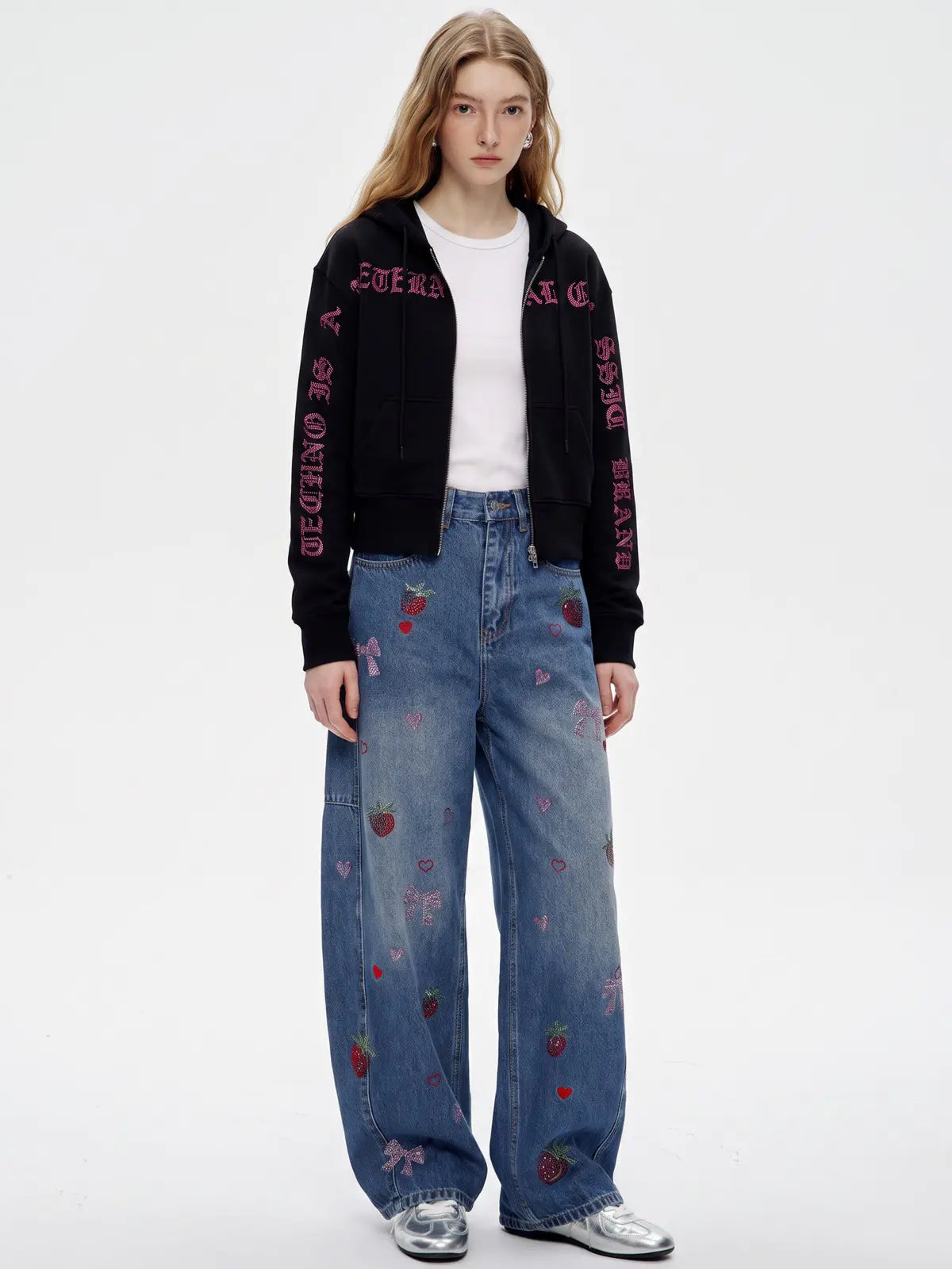 A model stands against a plain white studio background wearing a black cropped zip-up jacket with pink embroidered text down the sleeves, a white crew-neck T-shirt, and wide-leg blue jeans decorated with small pink and red strawberry patches. She is wearing silver sneakers. TCH Crystal Eagle Graphic Zip Up Hoodie