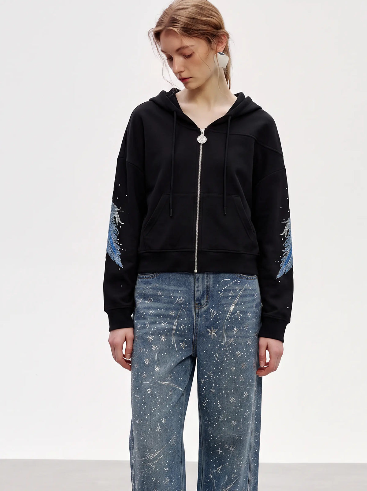 A model wearing a black zip-up hoodie with blue and white celestial print details on the sleeves, paired with blue jeans featuring a white starry pattern, against a plain light gray studio background. TCH Crystal Wing Graphic Zip Up Hoodie