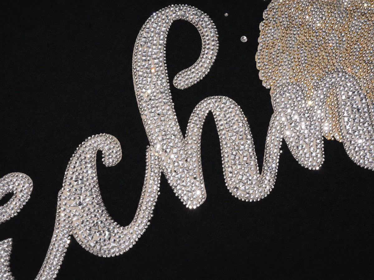 A close-up of a black garment featuring a large script word embellished with clear rhinestones in silver, alongside a textured gold embellishment. TCH Crystal Wing Graphic Zip Up Hoodie