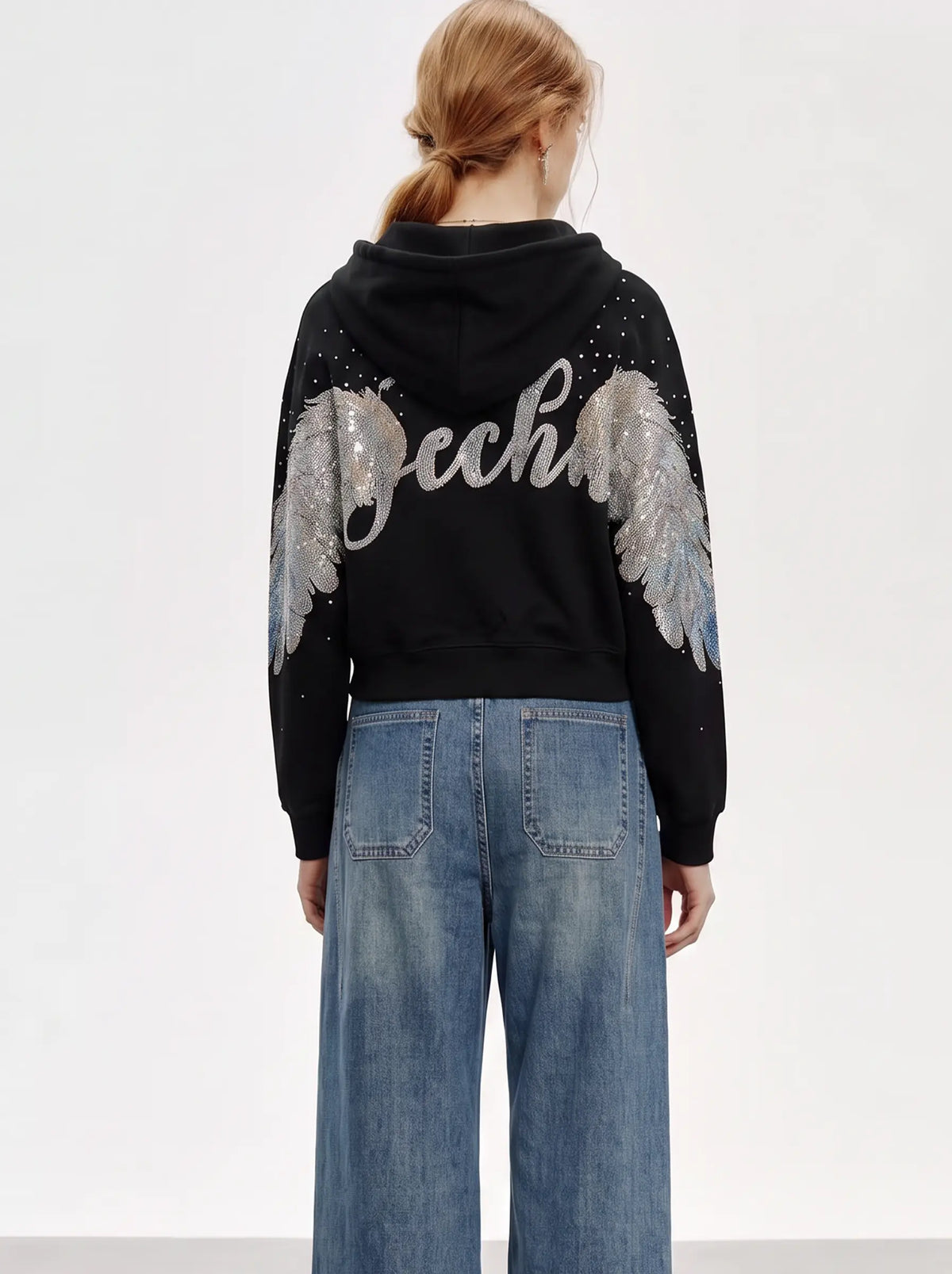 A model viewed from the back wearing a black cropped hoodie with large silver and white wing embellishments and sequins on the back, paired with wide-leg blue jeans against a plain white studio background. TCH Crystal Wing Graphic Zip Up Hoodie