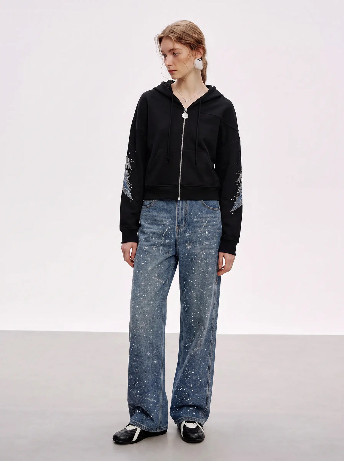 A model stands in a studio wearing a black cropped zip-up hoodie with silver sleeve details, blue wide-leg jeans, and black-and-white sneakers. TCH Crystal Wing Graphic Zip Up Hoodie