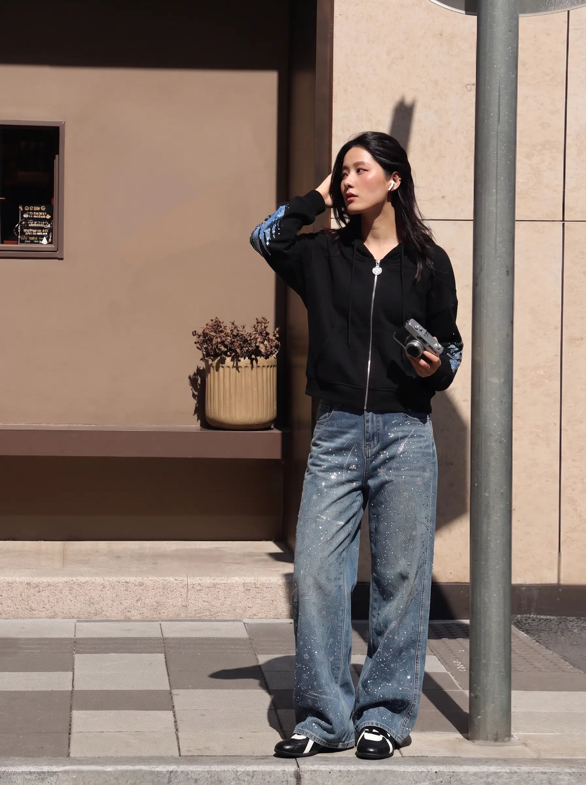 A woman stands outdoors on a sidewalk next to a building, wearing a black zip-up hoodie with blue patterned sleeves, wide-leg light-wash jeans, and black sneakers. TCH Crystal Wing Graphic Zip Up Hoodie