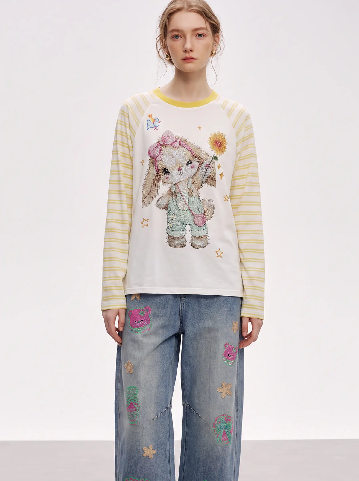 A model wearing a cream-colored long-sleeve t-shirt with yellow stripes on the sleeves, featuring a large cartoon bunny graphic on the front, paired with light blue jeans decorated with colorful patches. TCH Cute Bunny Rhinestone Graphic Striped Sleeve Long Sleeve T-Shirt