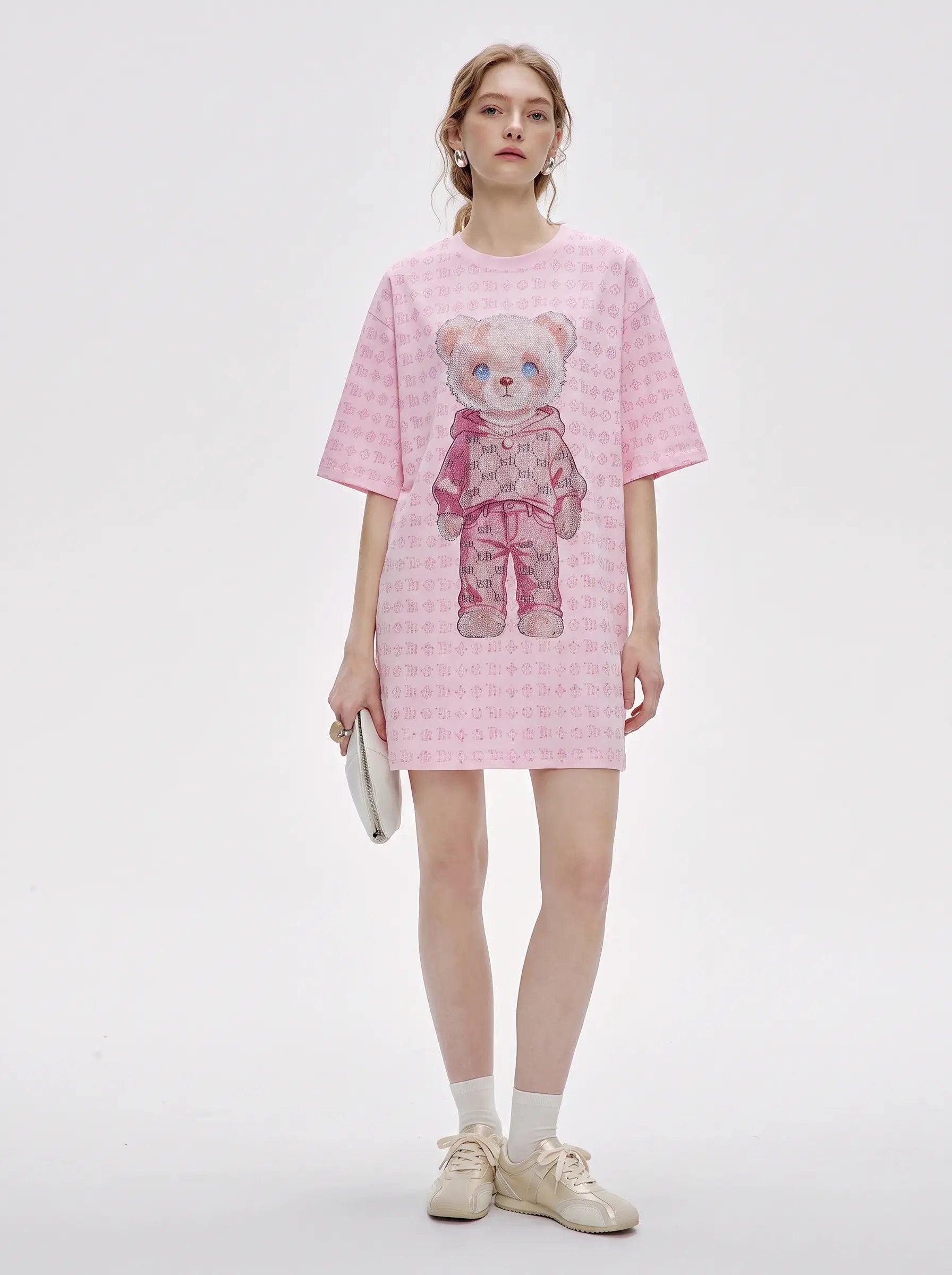 A model stands against a light gray studio background wearing a pink short-sleeve t-shirt dress with a repeating small pattern and a large teddy bear graphic on the front. She is holding a small white clutch and wearing white socks and beige sneakers. TCH Cute Hoodie Teddy Bear Rhinestone Oversized T-Shirt Dress