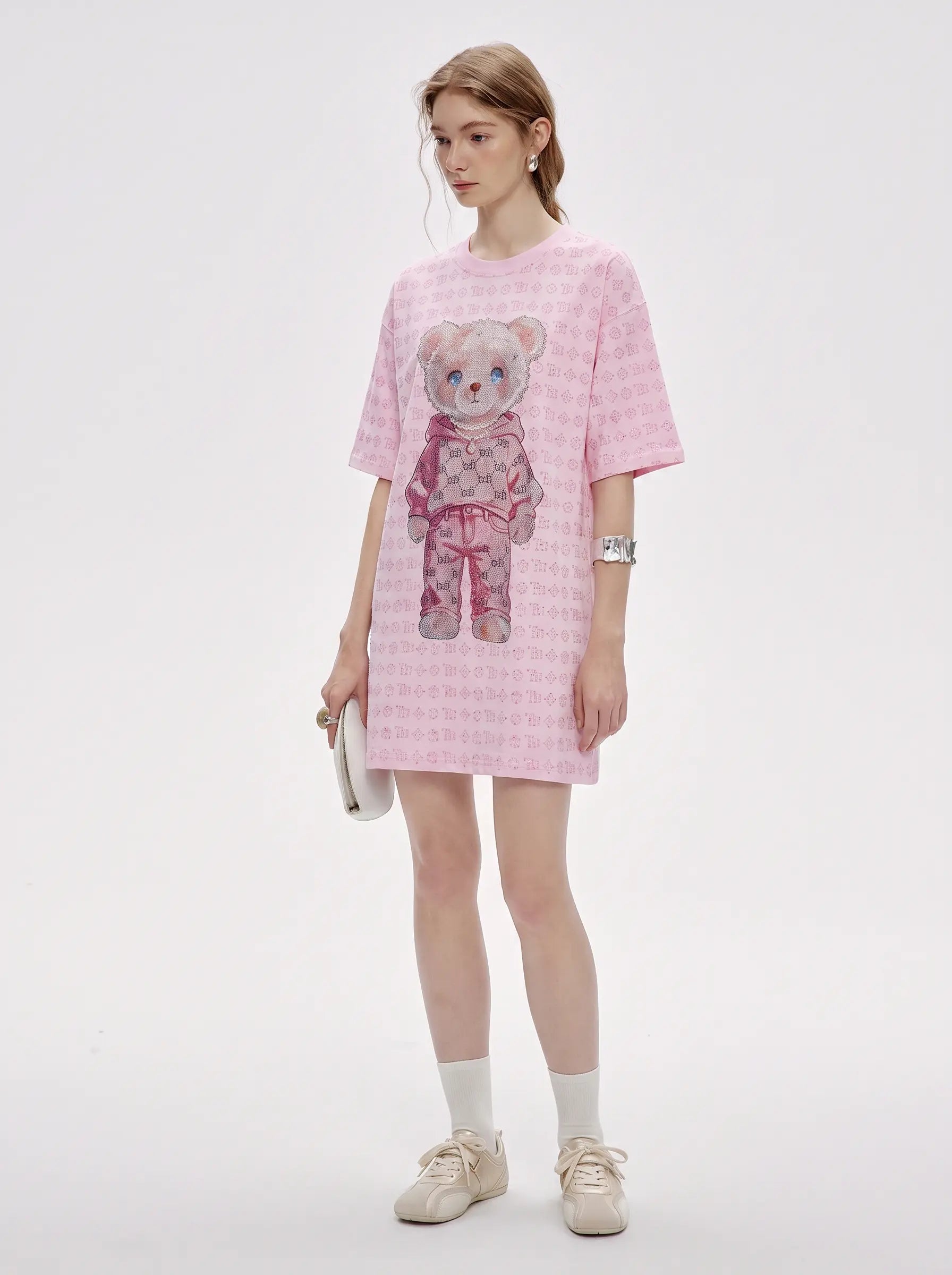 A model stands against a light gray studio background wearing a pink short-sleeve t-shirt dress with a repeating small pattern and a large teddy bear graphic on the front. She is styled with white socks, beige sneakers, and a small white clutch. TCH Cute Hoodie Teddy Bear Rhinestone Oversized T-Shirt Dress