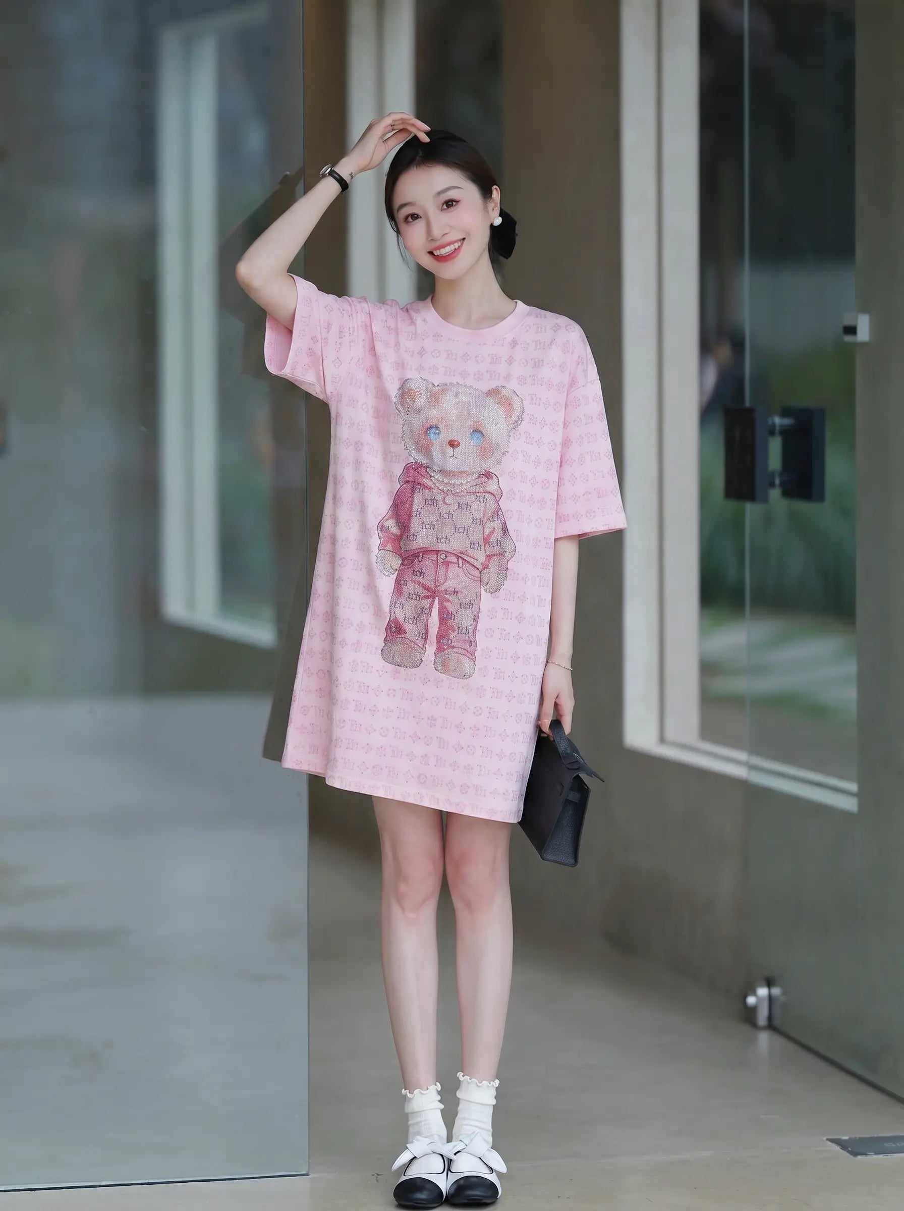 A woman modeling a pink gingham short-sleeve t-shirt dress with a teddy bear graphic on the front, paired with white socks and black-and-white sneakers, standing in a modern glass-walled corridor. TCH Cute Hoodie Teddy Bear Rhinestone Oversized T-Shirt Dress