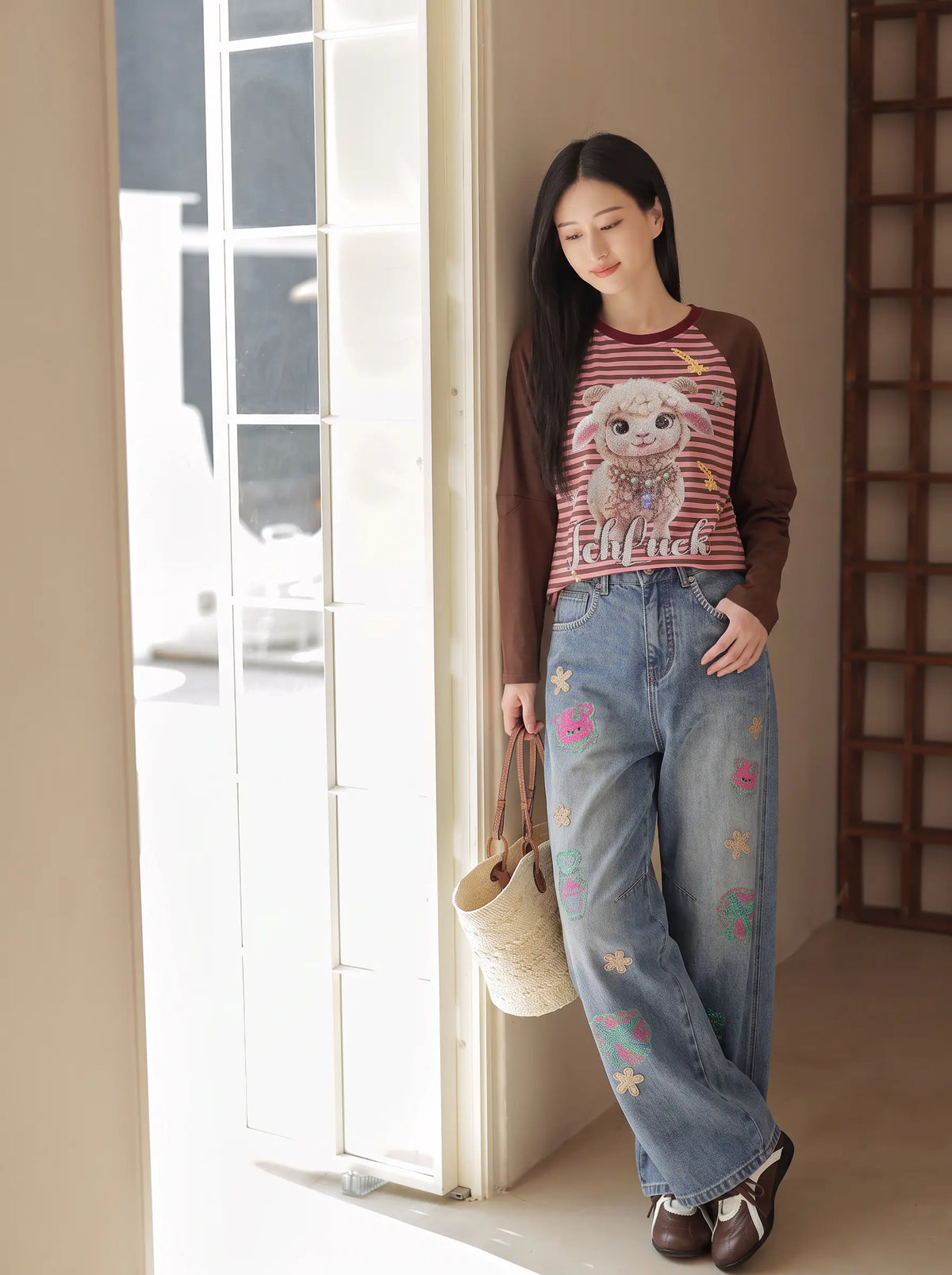 A woman stands indoors near a bright window, wearing a brown long-sleeve cardigan over a red-and-white striped graphic T-shirt with blue wide-leg jeans featuring colorful embroidered flowers and stars. She is holding a woven straw tote bag. TCH Cute Lamb Pink Striped Raglan Long Sleeve Top