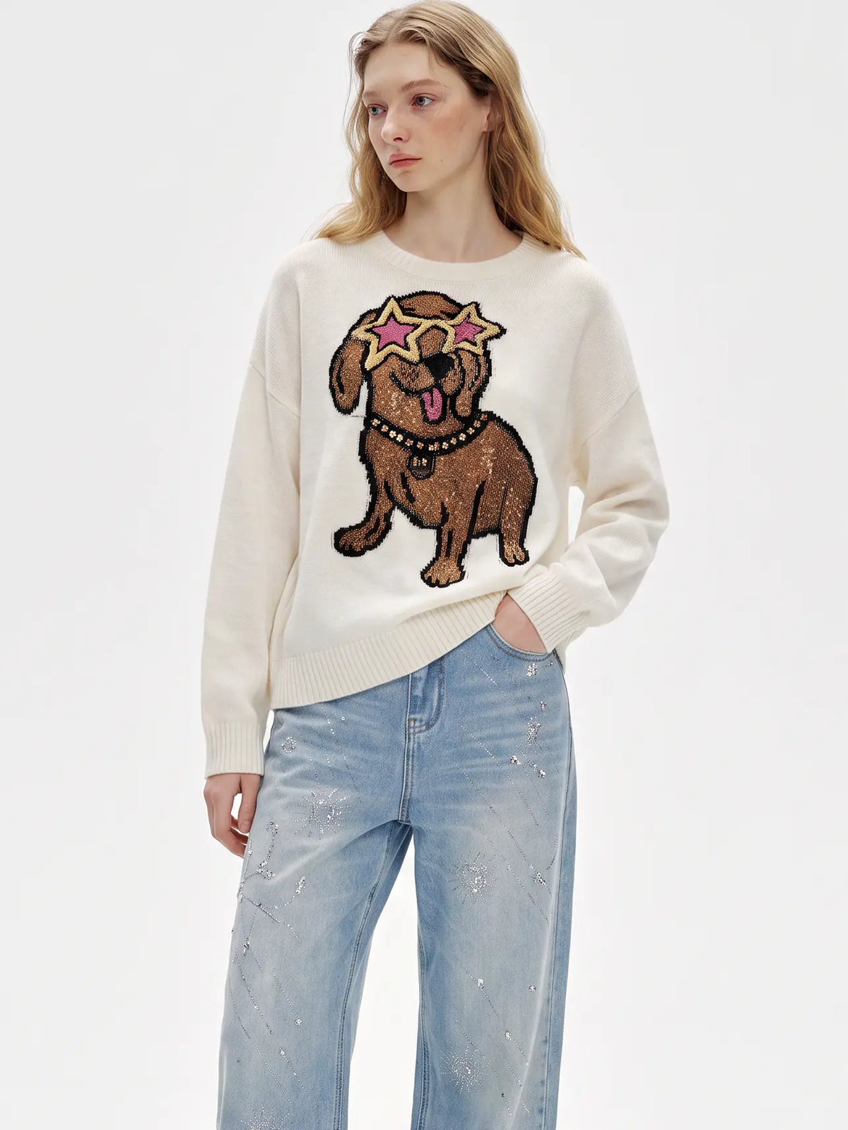 A woman modeling a cream-colored knit sweater featuring a large brown dog graphic with pink star-shaped sunglasses on the front, paired with light-wash blue jeans against a plain white studio background. TCH Cute Puppy Star Graphic Knit Sweater