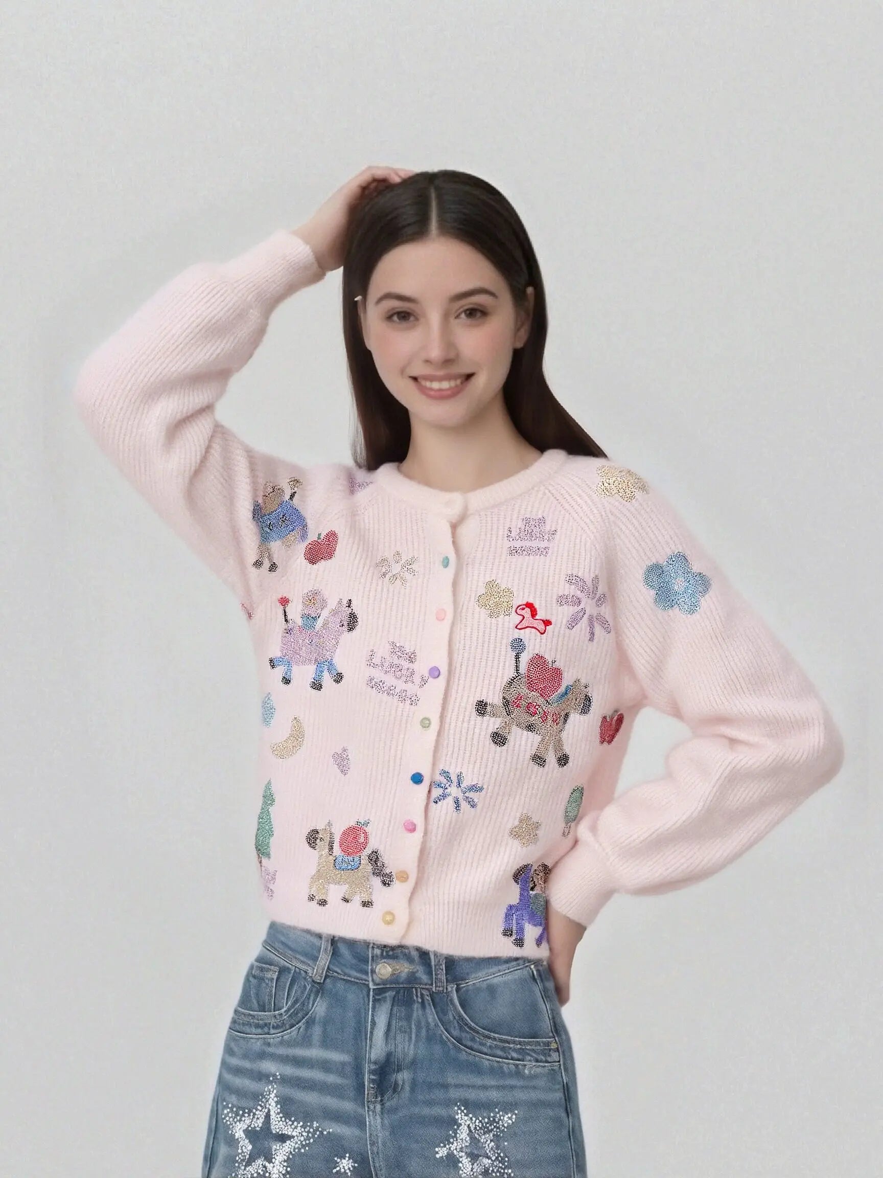 A woman wearing a light pink ribbed cardigan with colorful embroidered cartoon characters and blue jeans, posing against a light gray studio background. TCH Cute Rhinestone Animal Garden Knit Cardigan