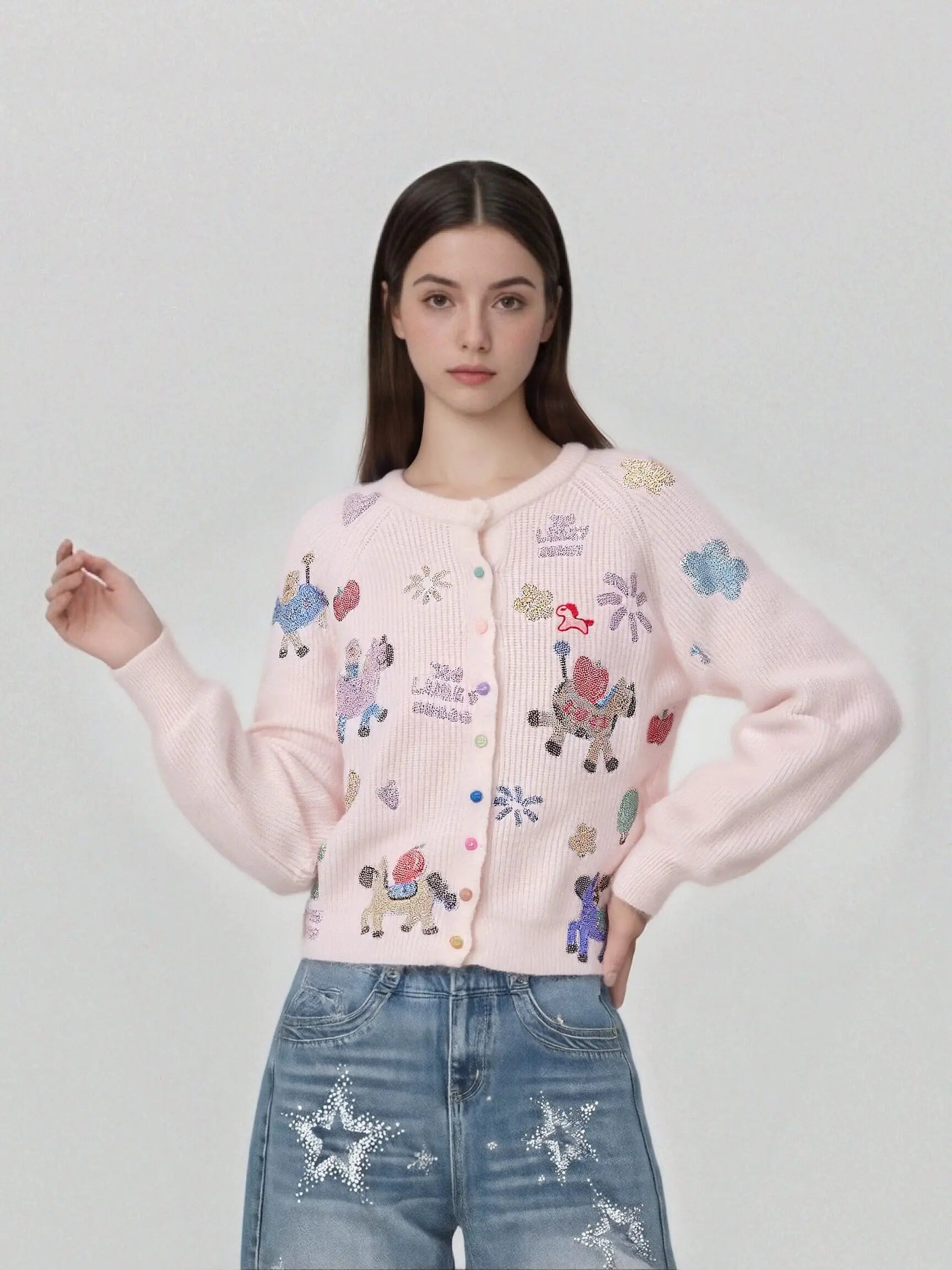 A woman modeling a light pink knit cardigan with colorful embroidered cartoon characters and text, paired with distressed blue jeans against a plain light gray studio background. TCH Cute Rhinestone Animal Garden Knit Cardigan
