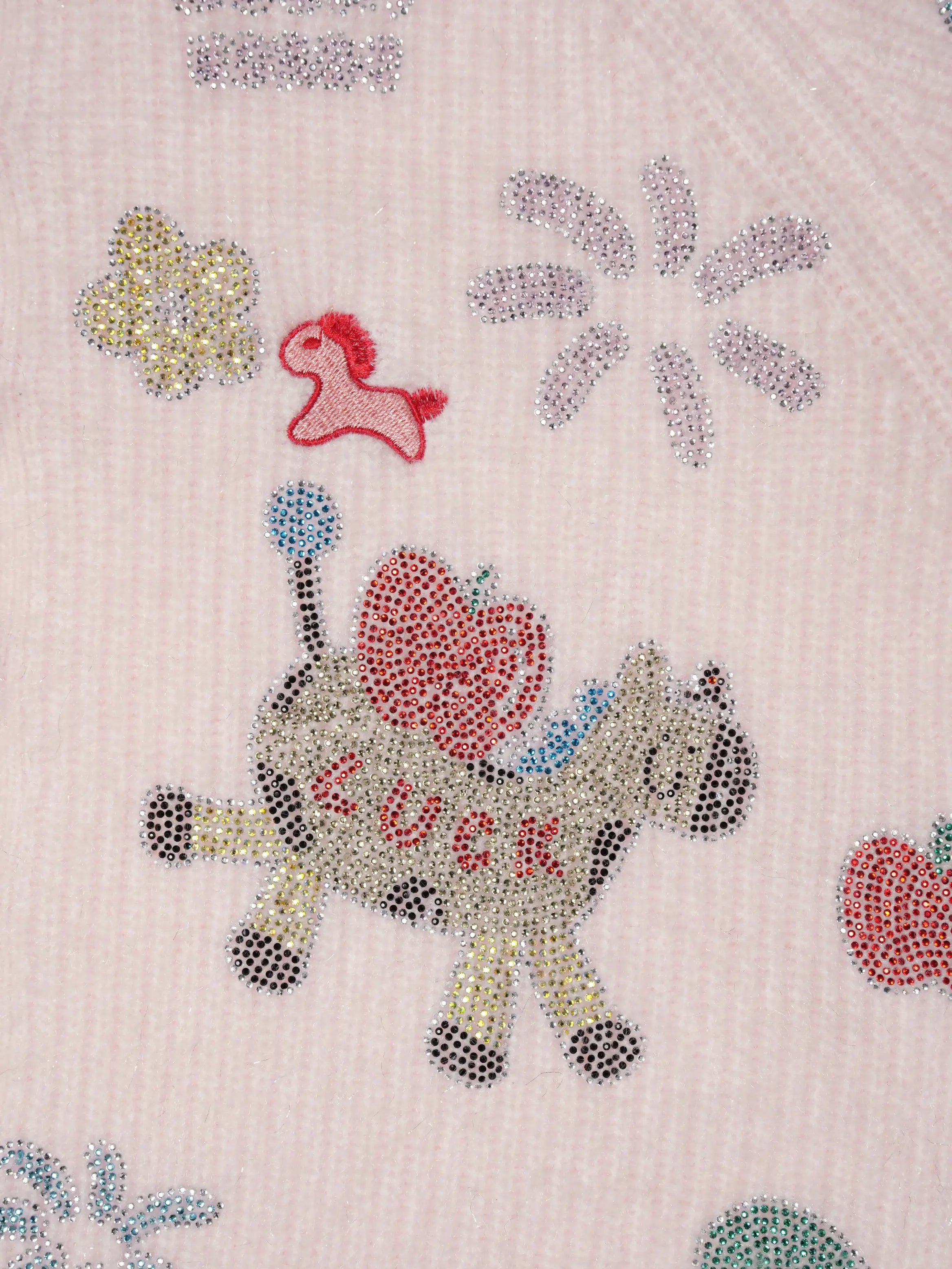 A close-up of a light pink textured fabric featuring a whimsical design of a cow decorated with rhinestones, a red heart-shaped apple, the word 'LUCK' in rhinestones, a small red rocking horse, and other scattered rhinestone motifs. TCH Cute Rhinestone Animal Garden Knit Cardigan