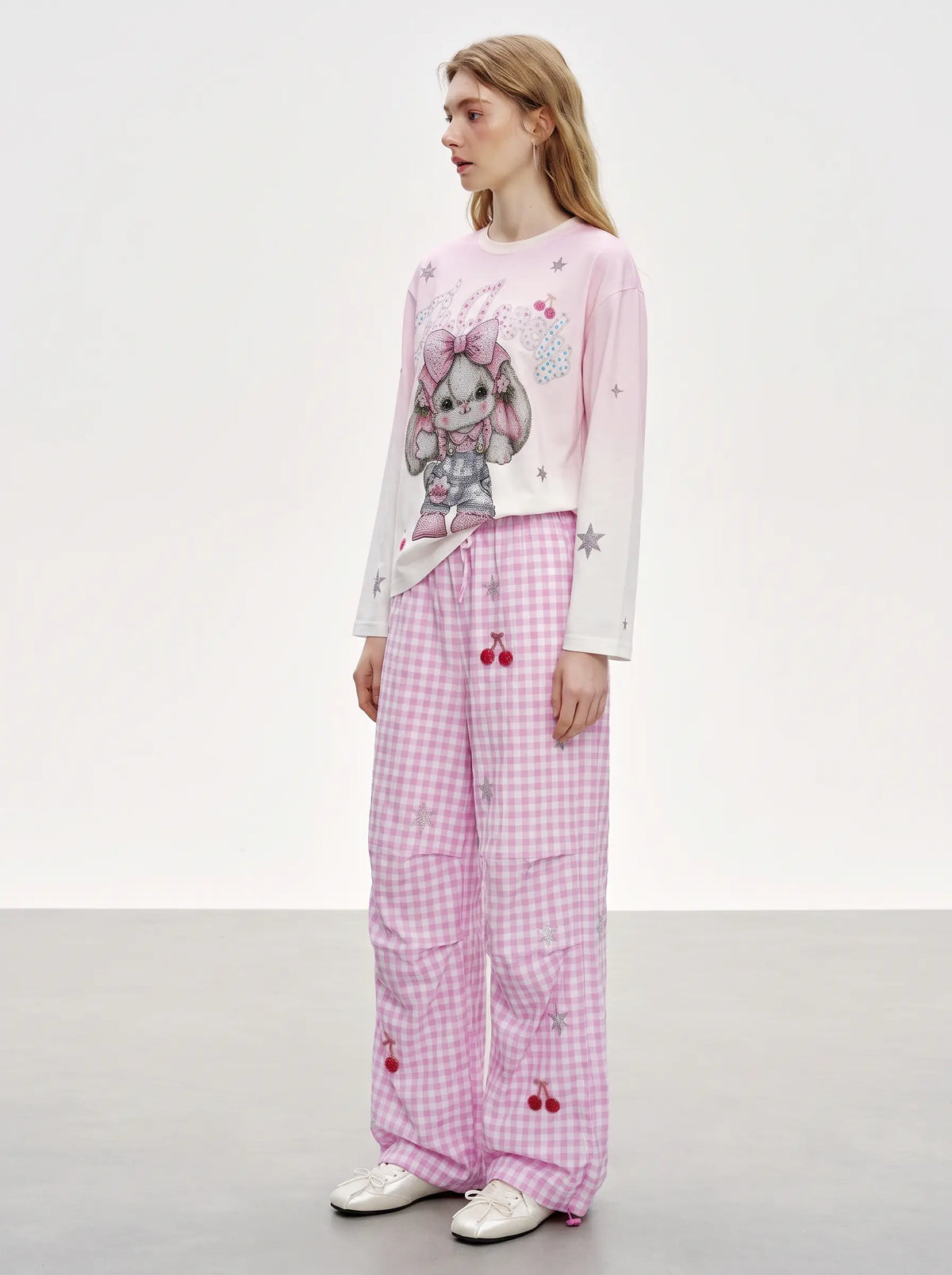 A model stands in a studio wearing a pink long-sleeve top with a cartoon bunny graphic and star details, paired with pink gingham pants featuring cherry motifs. TCH Cute Rhinestone Cat Graphic Pink Long Sleeve T-Shirt