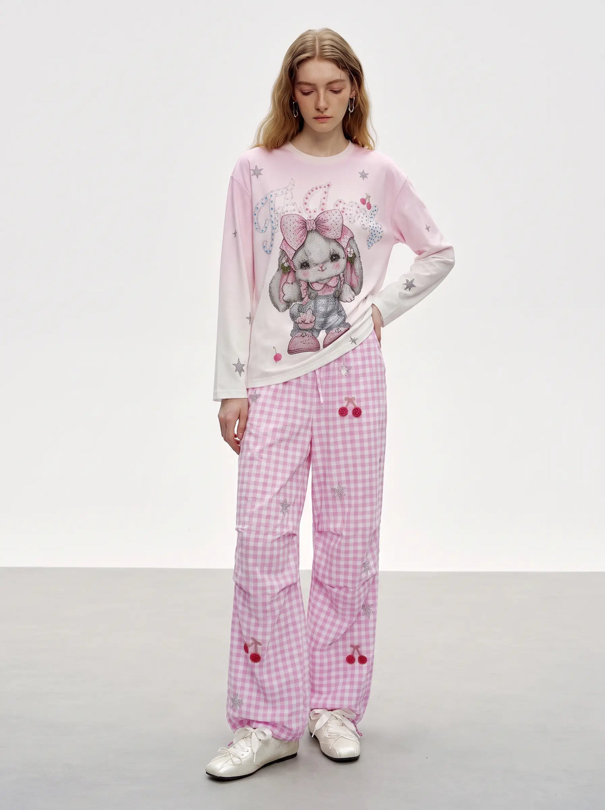 A model wearing a pink and white long-sleeve graphic t-shirt with a cartoon bunny design, paired with matching pink and white gingham pants featuring cherry details, against a plain studio background. TCH Cute Rhinestone Cat Graphic Pink Long Sleeve T-Shirt