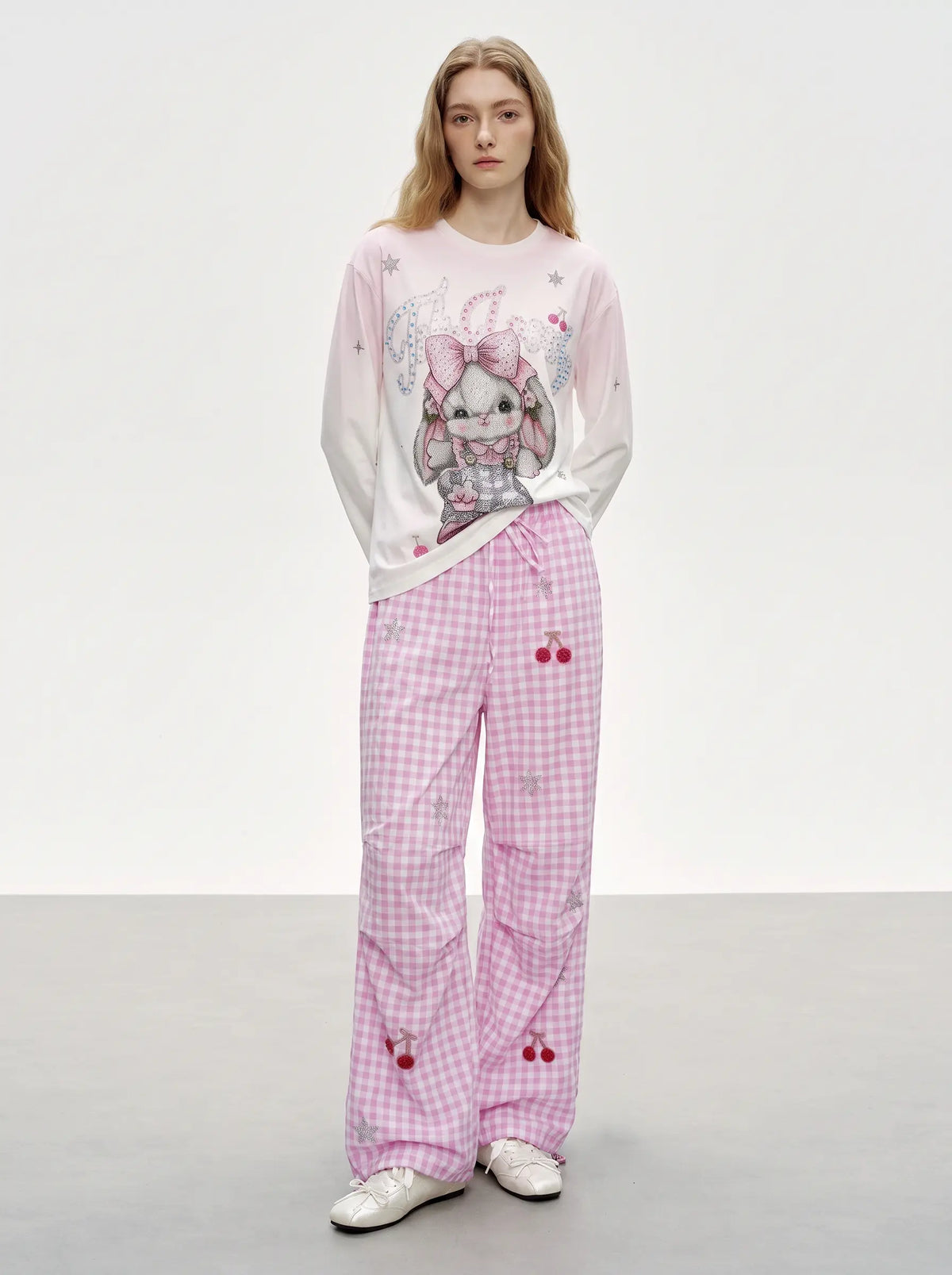 A model stands in a studio wearing a light pink and white long-sleeve top with a cartoon bunny graphic, paired with matching pink gingham pajama pants featuring cherry and bunny motifs, and white sneakers. TCH Cute Rhinestone Cat Graphic Pink Long Sleeve T-Shirt