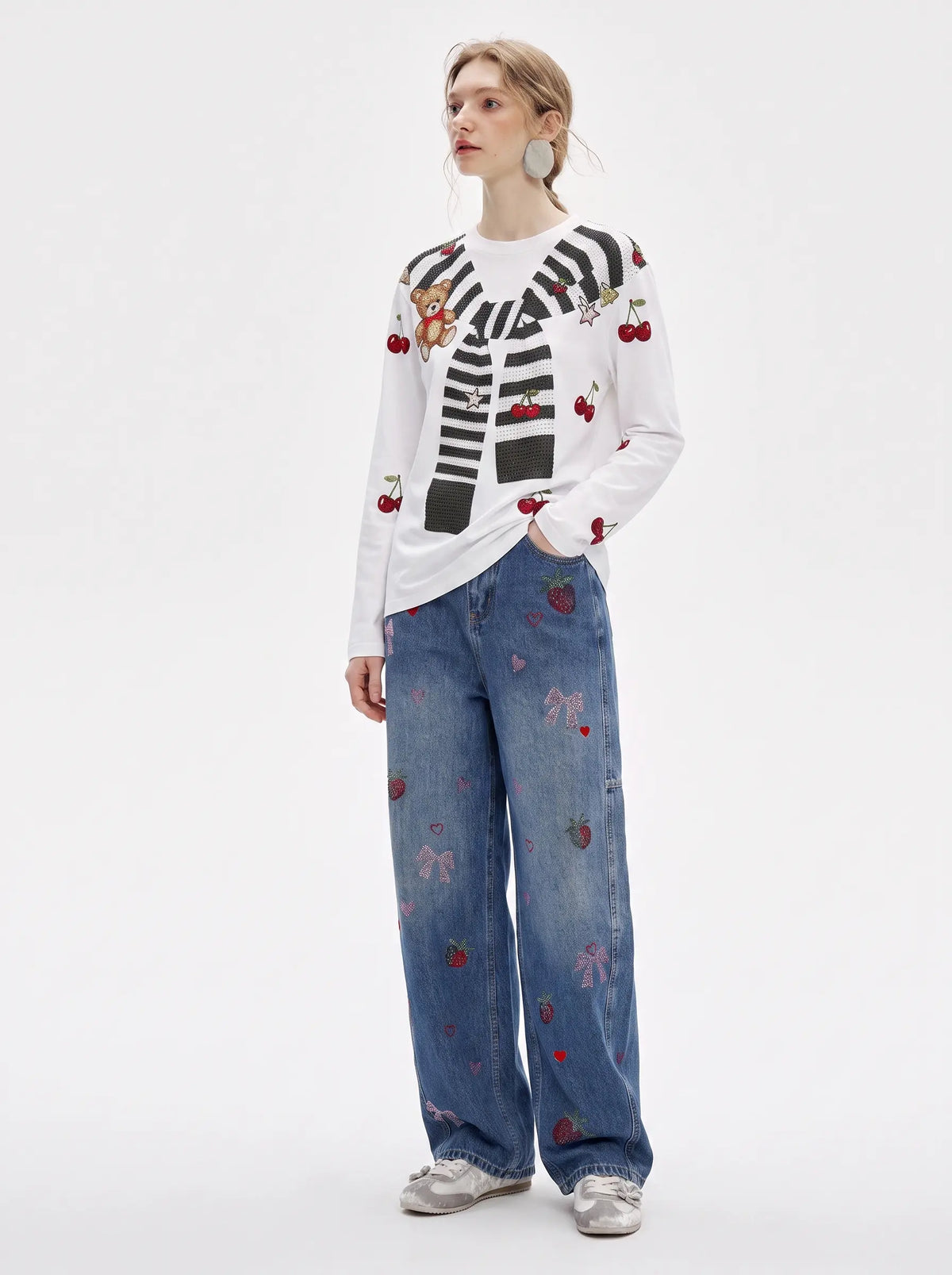 A model stands against a plain white studio background wearing a white long-sleeve top with black-and-white striped and cherry embroidery, blue wide-leg jeans with pink bow and red strawberry embroidery, and gray sneakers. TCH Cute Teddy Bear Cherry Print Long Sleeve Graphic T-Shirt