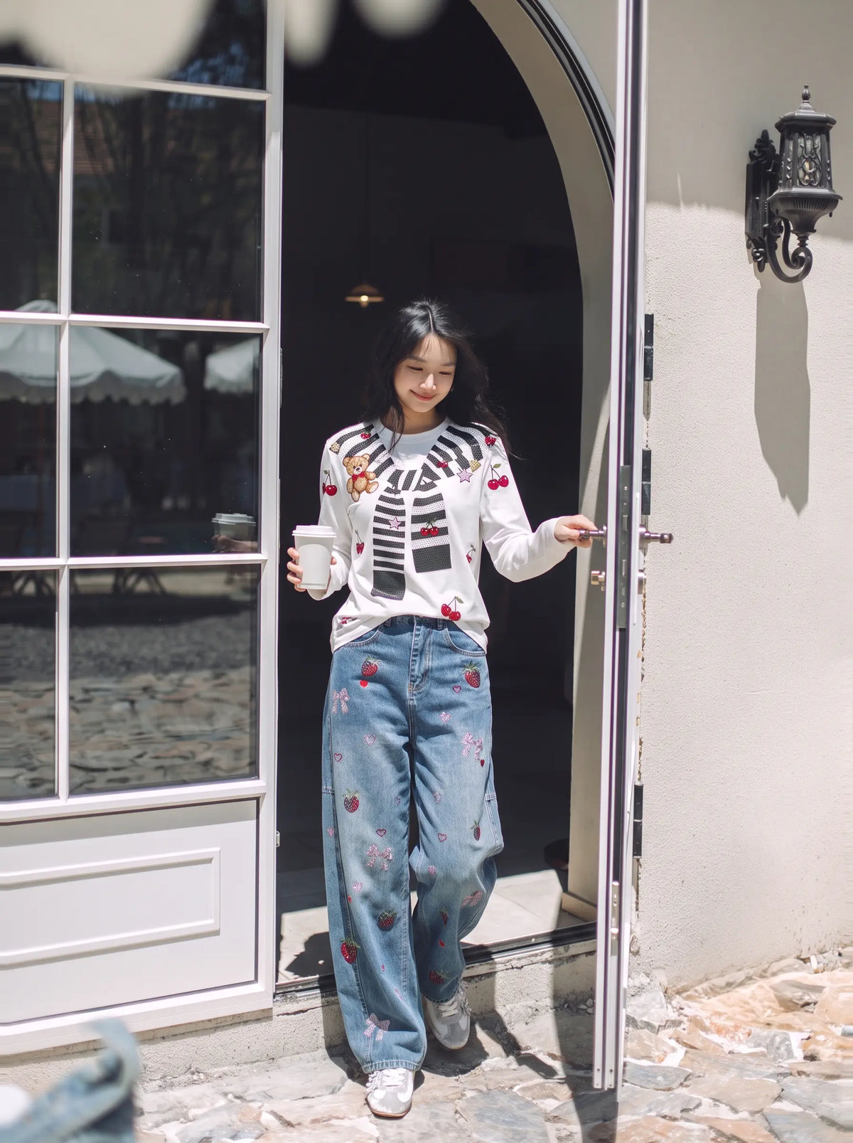 A woman stands in an open doorway wearing a white long-sleeve top with a black and red graphic print and loose blue jeans with red floral embroidery, holding a white coffee mug. TCH Cute Teddy Bear Cherry Print Long Sleeve Graphic T-Shirt