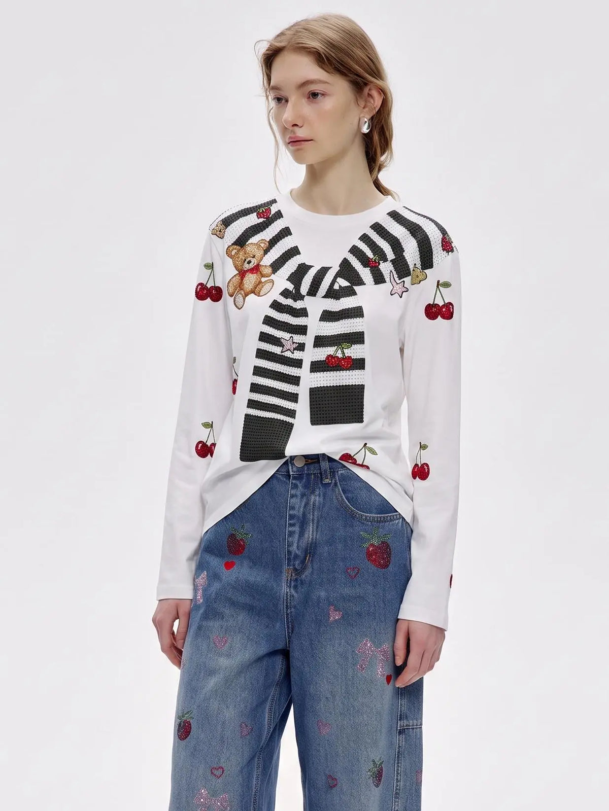 A model wearing a white long-sleeve knit sweater with black stripes, cherries, strawberries, and a teddy bear motif, paired with blue jeans featuring strawberry prints, against a plain white studio background. TCH Cute Teddy Bear Cherry Print Long Sleeve Graphic T-Shirt
