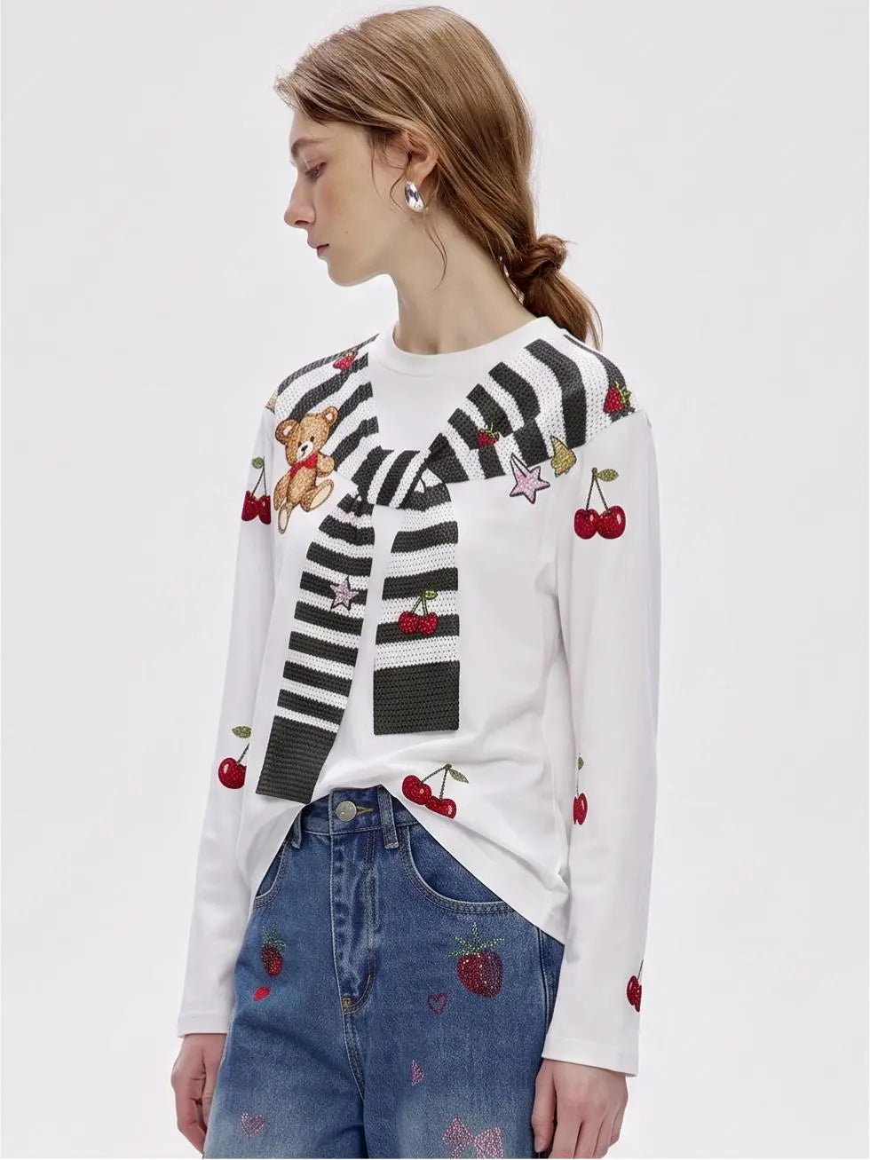 A woman wearing a white long-sleeve knit sweater with black-and-white striped yoke details, embroidered cherries, a teddy bear, and stars, paired with blue jeans featuring strawberry embroidery, against a plain light gray studio background. TCH Cute Teddy Bear Cherry Print Long Sleeve Graphic T-Shirt