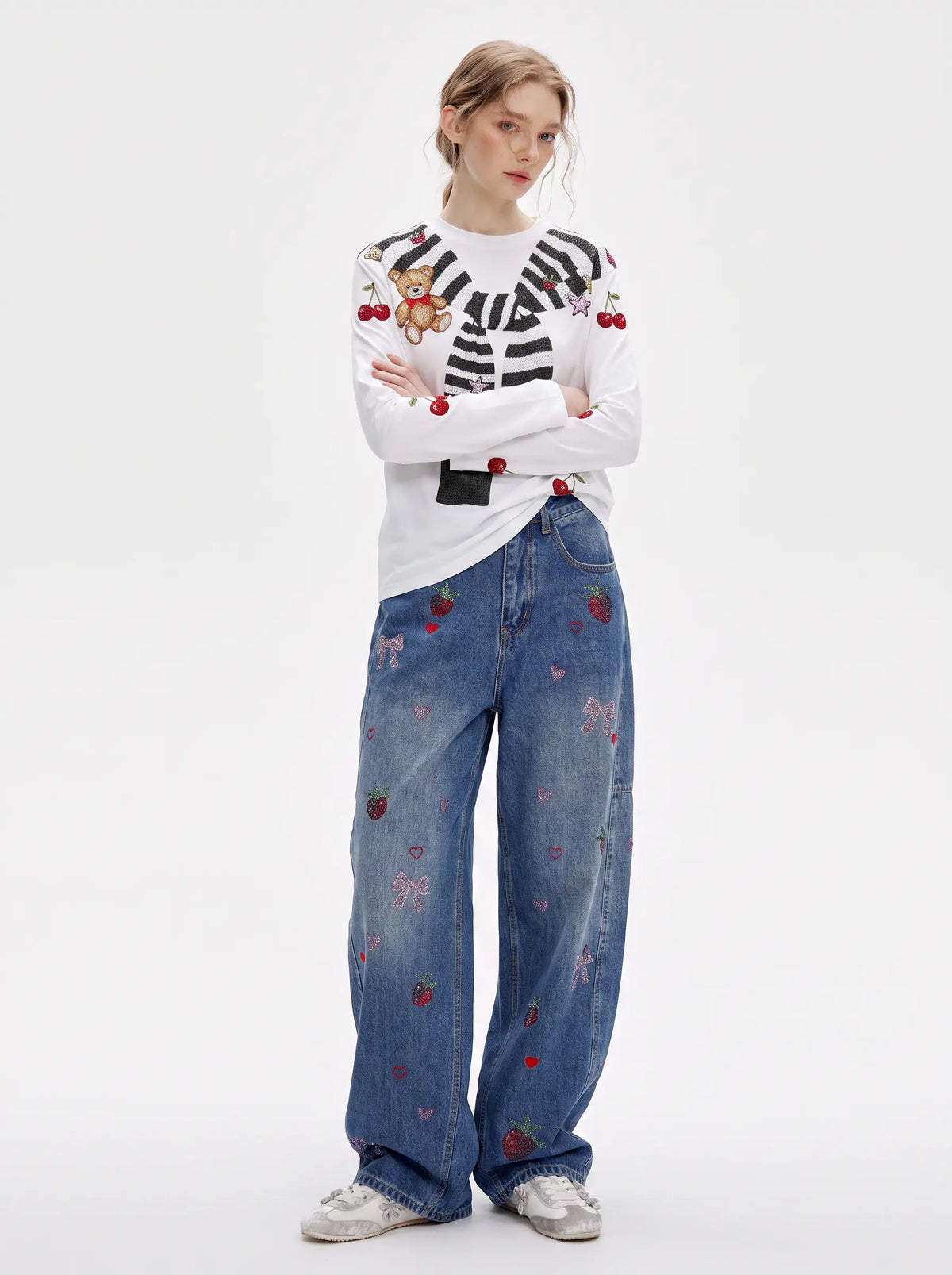 A model stands against a light gray studio background wearing a white long-sleeve top with black-and-white striped detailing, embroidered cherries, and a teddy bear motif on the chest, paired with wide-leg blue jeans featuring scattered red embroidered strawberries and hearts, and white sneakers. TCH Cute Teddy Bear Cherry Print Long Sleeve Graphic T-Shirt