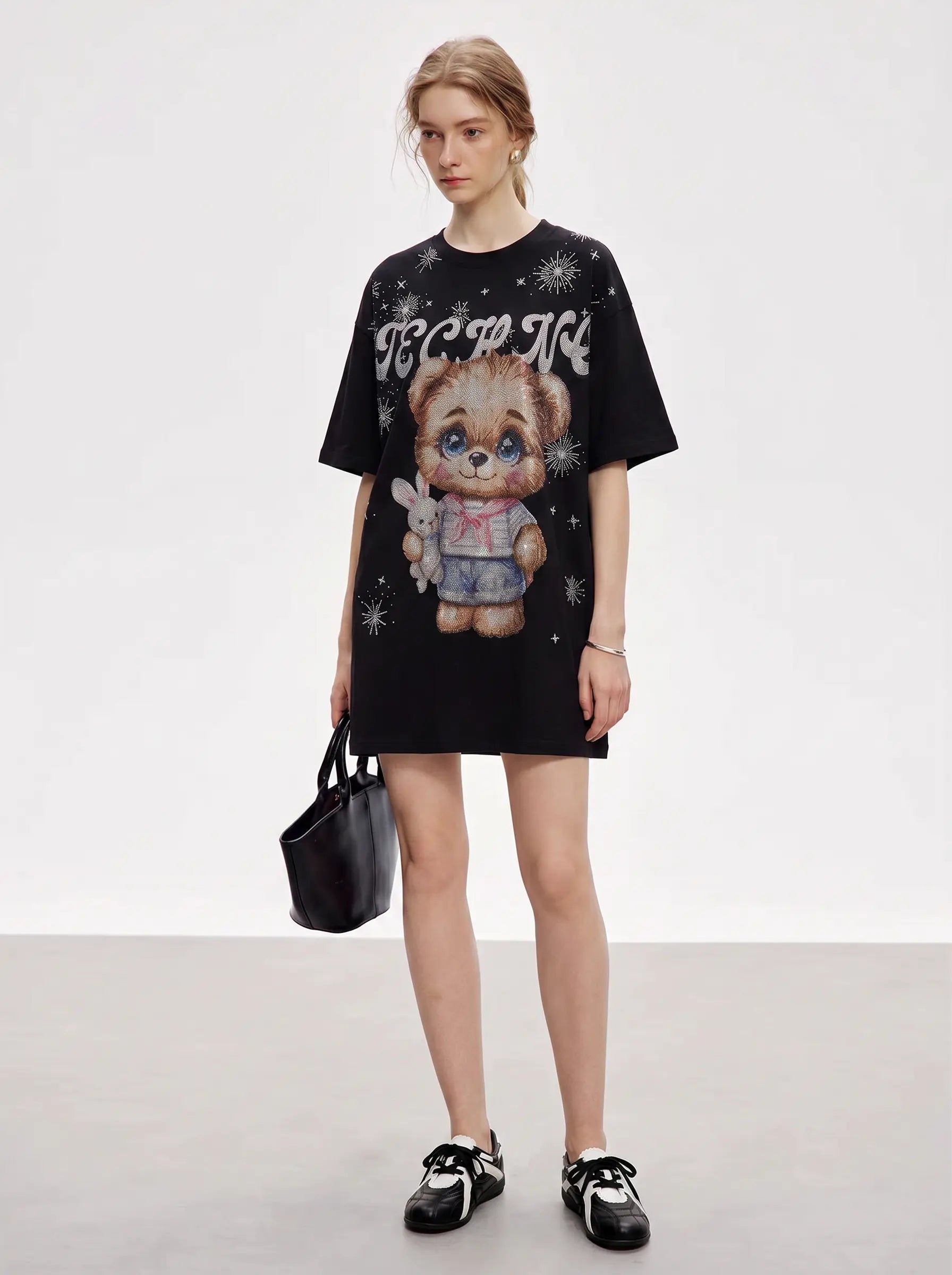 A model stands in a studio wearing an oversized black graphic T-shirt dress with a teddy bear illustration and white gothic-style text, paired with black-and-white sneakers and holding a black handbag. TCH Cute Teddy Bear Rhinestone Oversized T-Shirt Dress