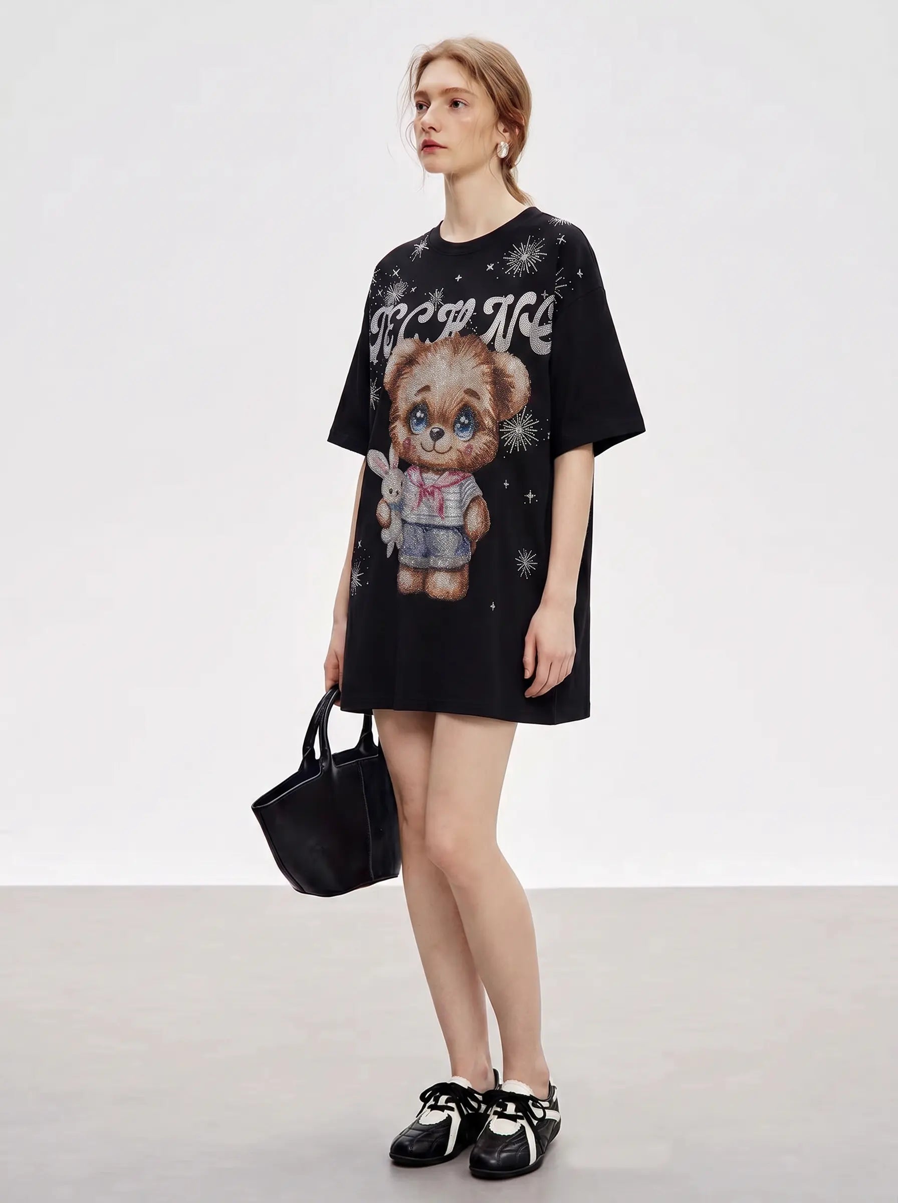 A model stands in a studio wearing an oversized black graphic T-shirt dress with a teddy bear illustration and white text, holding a black handbag and wearing black Mary Jane shoes. TCH Cute Teddy Bear Rhinestone Oversized T-Shirt Dress