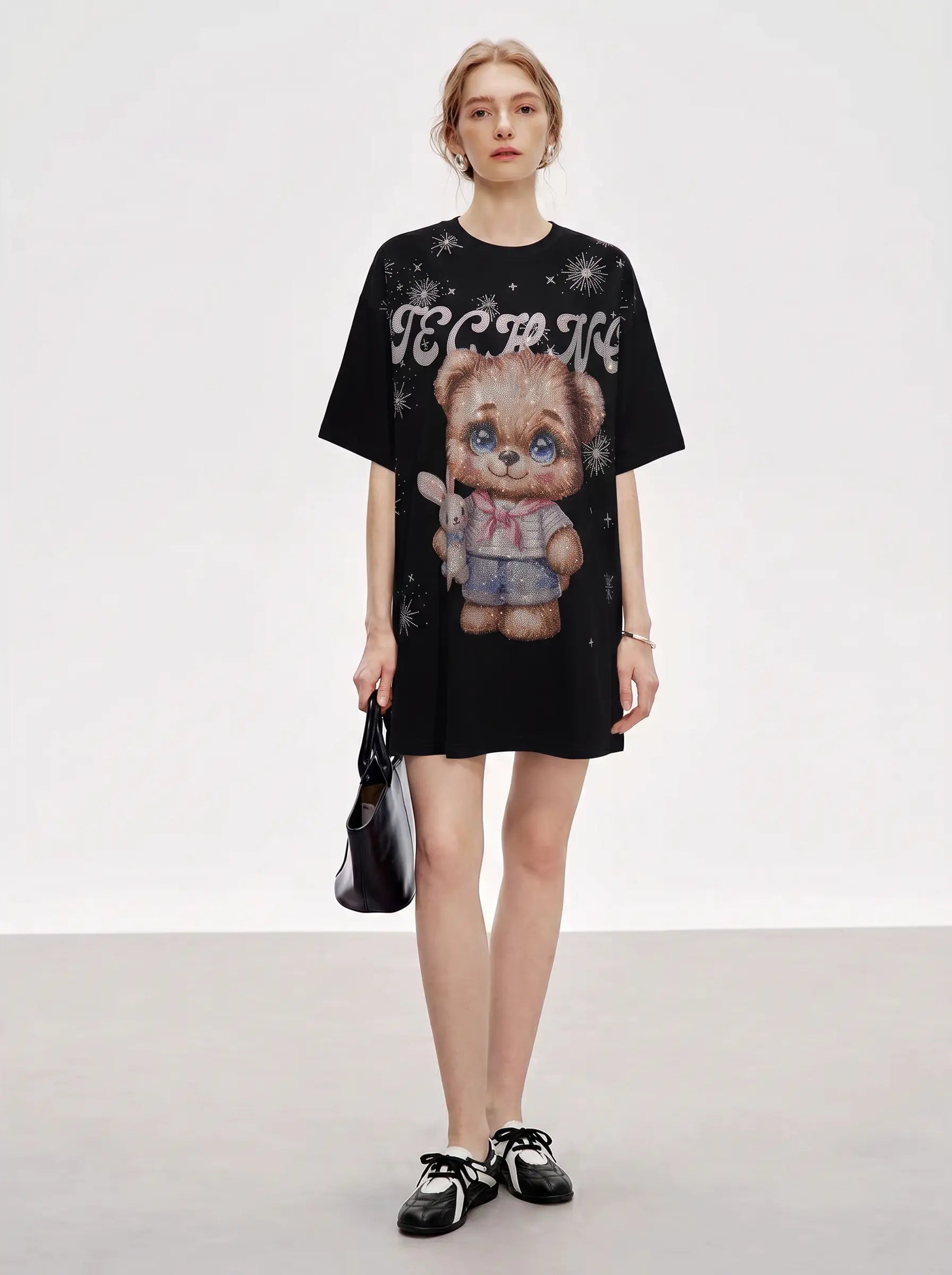 A model stands against a light gray studio background wearing an oversized black graphic T-shirt dress with a teddy bear print and white gothic lettering, paired with black-and-white sneakers and holding a black handbag. TCH Cute Teddy Bear Rhinestone Oversized T-Shirt Dress