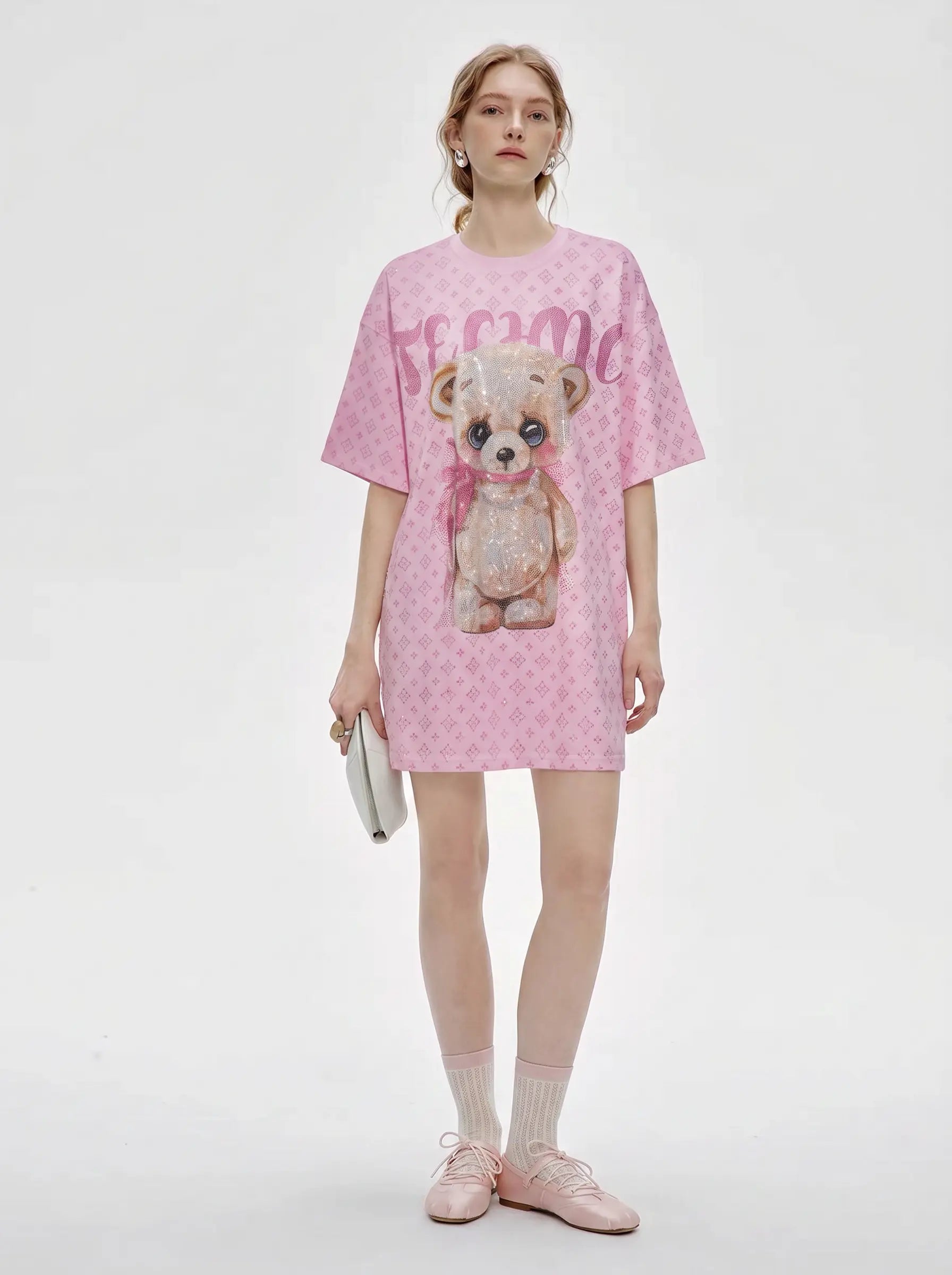 A model stands against a light gray studio background wearing a pink short-sleeve T-shirt dress with a teddy bear graphic and the word 'tomoe' across the front, paired with white socks and pink shoes. TCH Cute Teddy Bear Rhinestone Oversized T-Shirt Dress