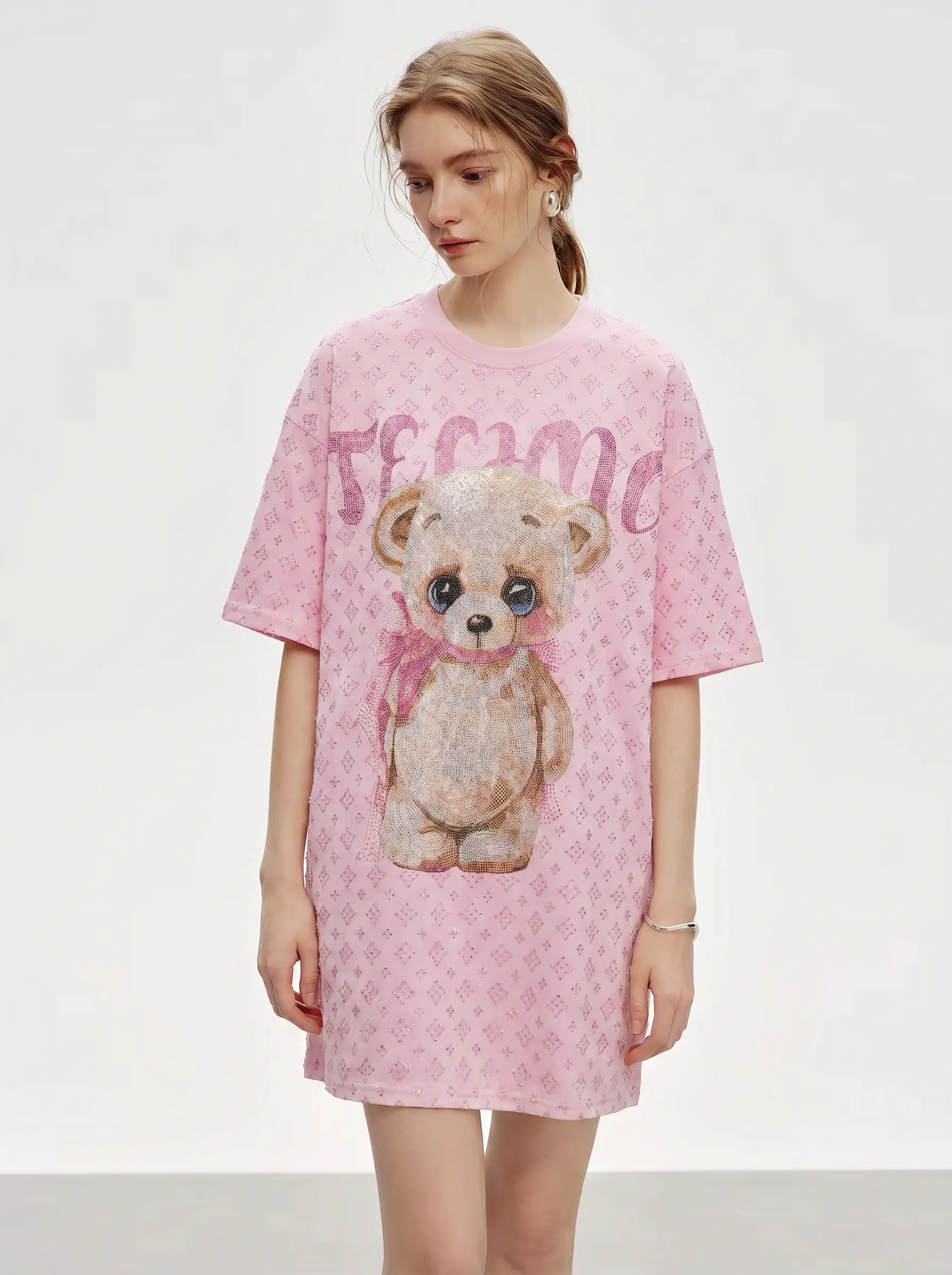 A woman modeling a pink short-sleeve t-shirt dress with a teddy bear graphic and monogram pattern, set against a plain light gray studio background. TCH Cute Teddy Bear Rhinestone Oversized T-Shirt Dress