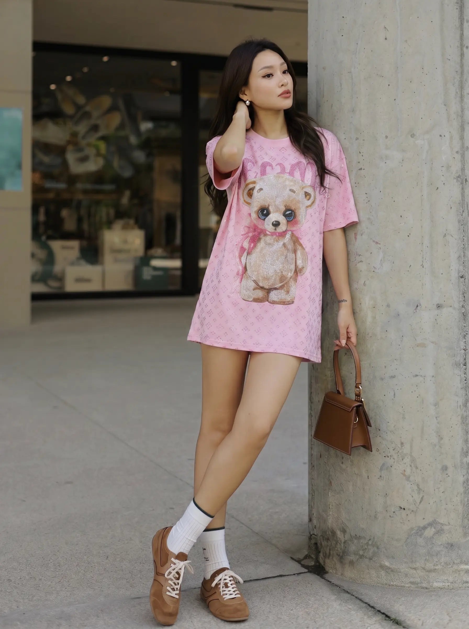 A woman stands outdoors leaning against a concrete pillar, wearing a pink oversized graphic T-shirt dress with a teddy bear print, white socks, brown lace-up shoes, and carrying a small brown handbag. TCH Cute Teddy Bear Rhinestone Oversized T-Shirt Dress