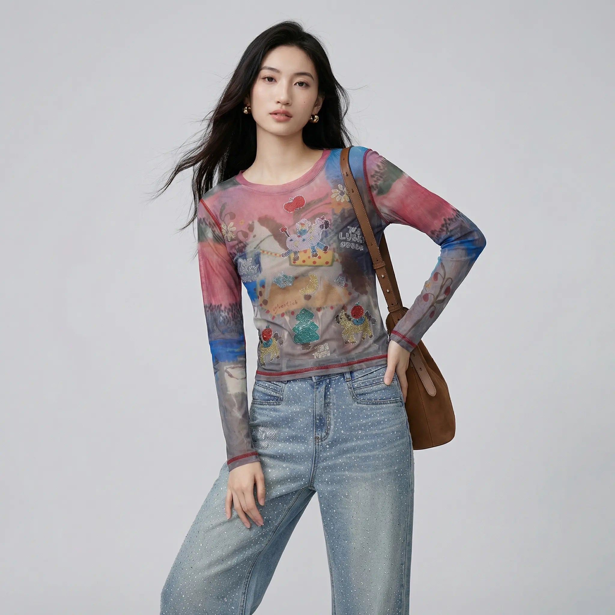 A woman modeling a long-sleeve sheer mesh top with a colorful cartoon and animal print, paired with light blue jeans and a brown shoulder bag, against a plain light gray studio background. TCH Dream Garden Rhinestone Mesh Long Sleeve Top