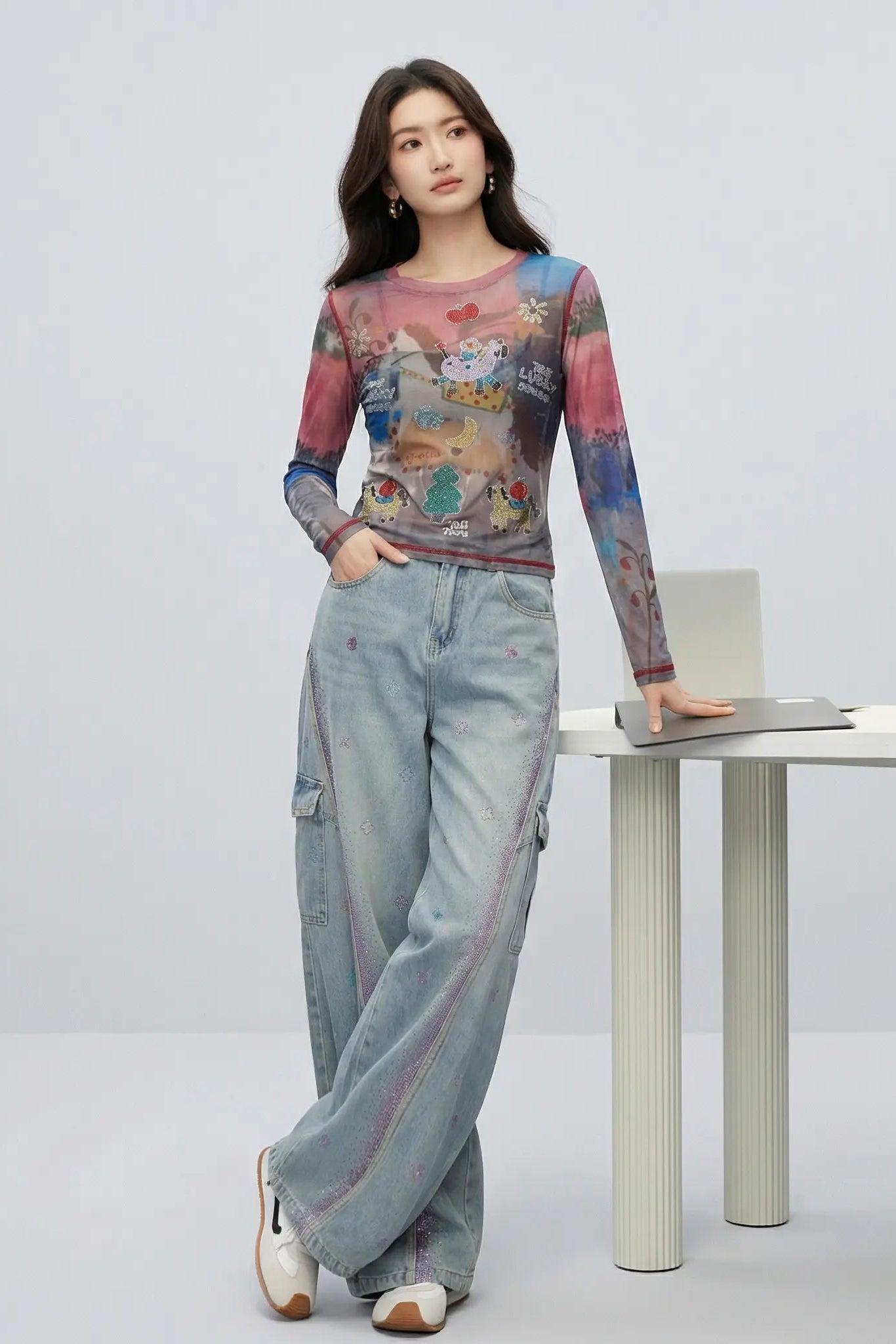 A woman stands in a studio wearing a long-sleeve sheer mesh top with a colorful graffiti-style print and light blue wide-leg cargo pants, posed beside a white table with a laptop. TCH Dream Garden Rhinestone Mesh Long Sleeve Top