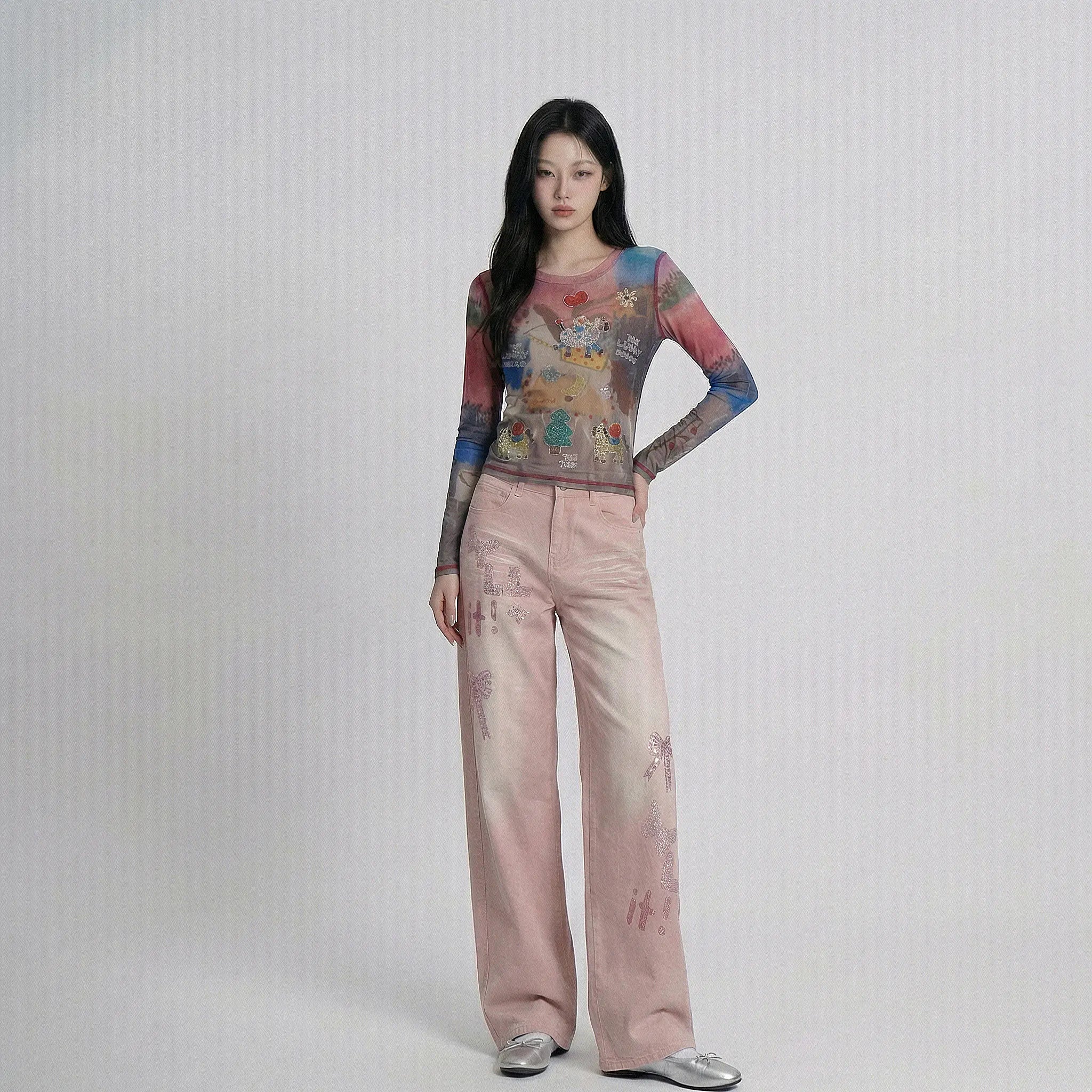 A model stands against a light gray studio background wearing a sheer long-sleeve top with a colorful abstract print and wide-leg pink pants with subtle tonal floral prints, paired with silver shoes. TCH Dream Garden Rhinestone Mesh Long Sleeve Top