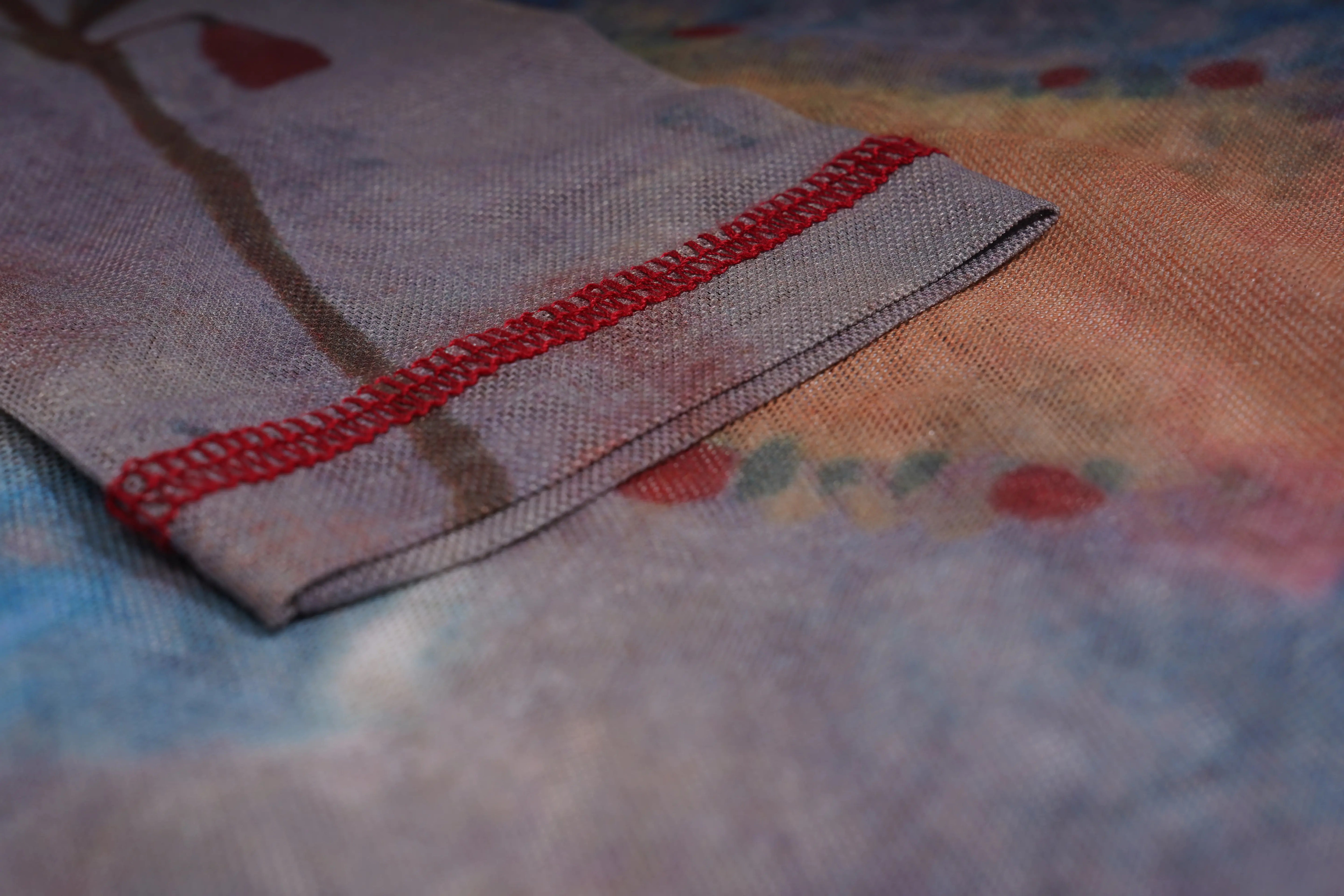 A close-up of a folded piece of textured fabric with a red stitched border, set against a soft, blurred multicolored fabric background. TCH Dream Garden Rhinestone Mesh Long Sleeve Top