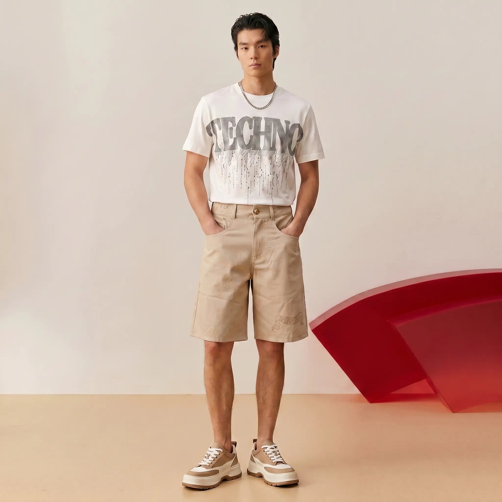 A man stands in a studio wearing a white graphic T-shirt with large gray 'TECHNO' lettering, beige shorts, and beige-and-white sneakers. A red sculptural form is visible in the background. TCH Dripping Rhinestone Logo Graphic T-Shirt