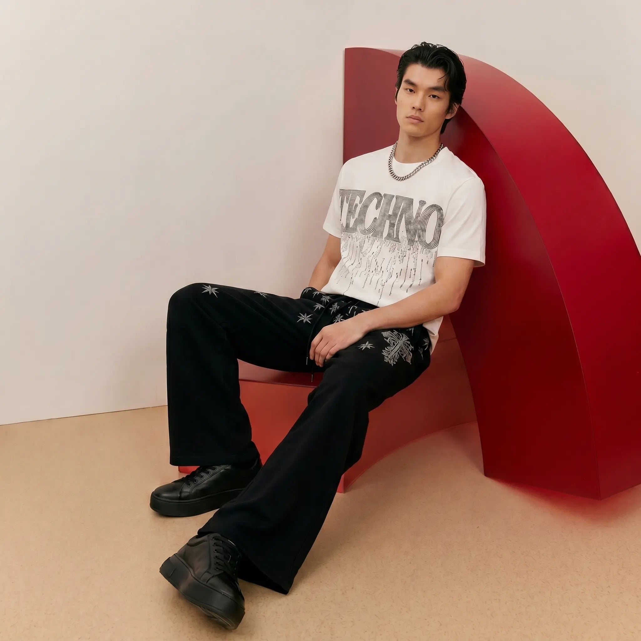 A man sits on a bold red curved chair in a minimalist studio setting. He is wearing a white graphic T-shirt with 'TECHNO' and a distressed graphic print, black wide-leg trousers with silver embroidered details, and black leather sneakers. TCH Dripping Rhinestone Logo Graphic T-Shirt