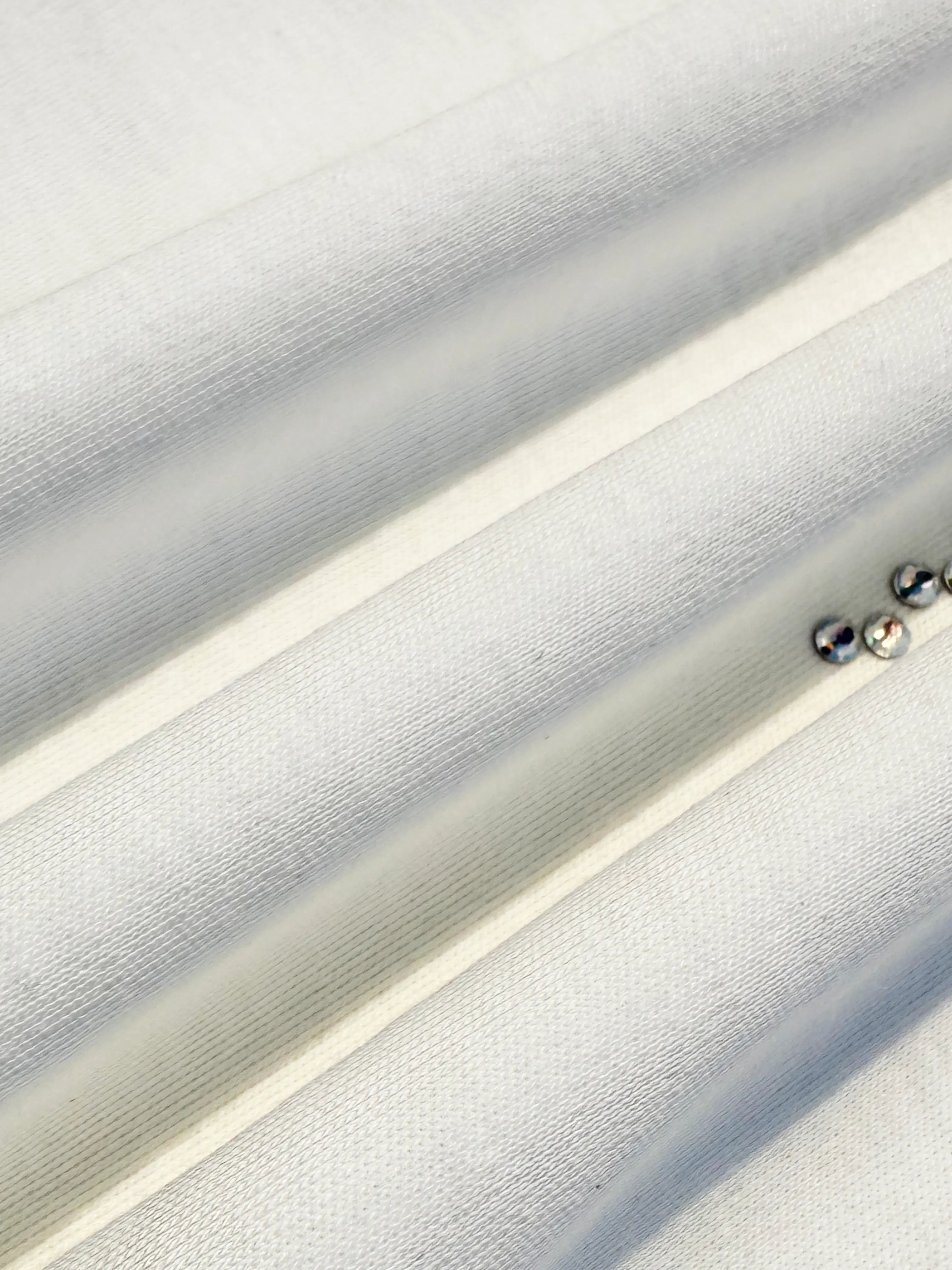 A close-up of a light gray or off-white textured fabric with diagonal folds and two small metallic studs on the right side. TCH Dripping Rhinestone Logo Graphic T-Shirt