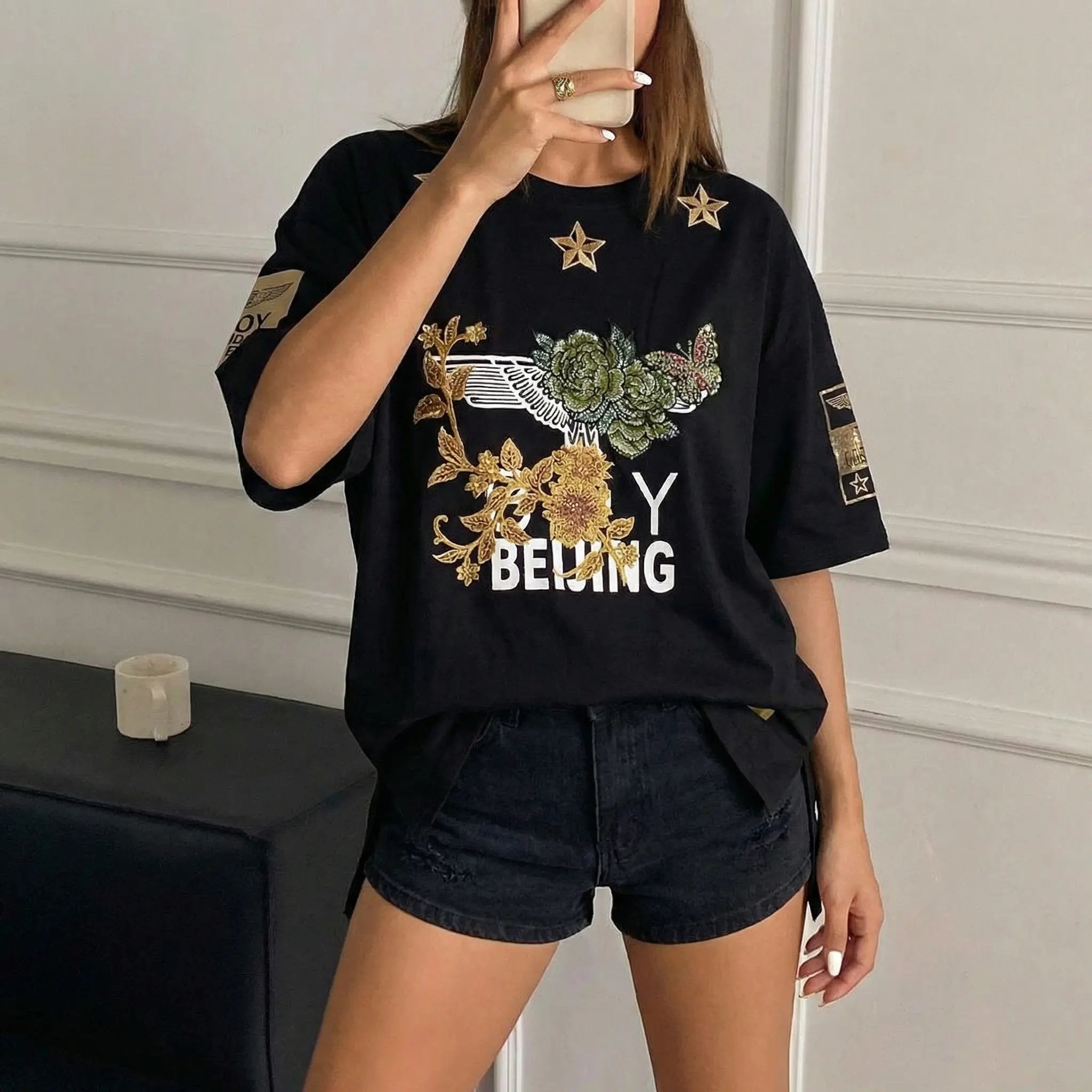 Person wearing a black t-shirt with floral design and text in an indoor setting, TCH Embroidered Floral Butterfly Rhinestone T-Shirt