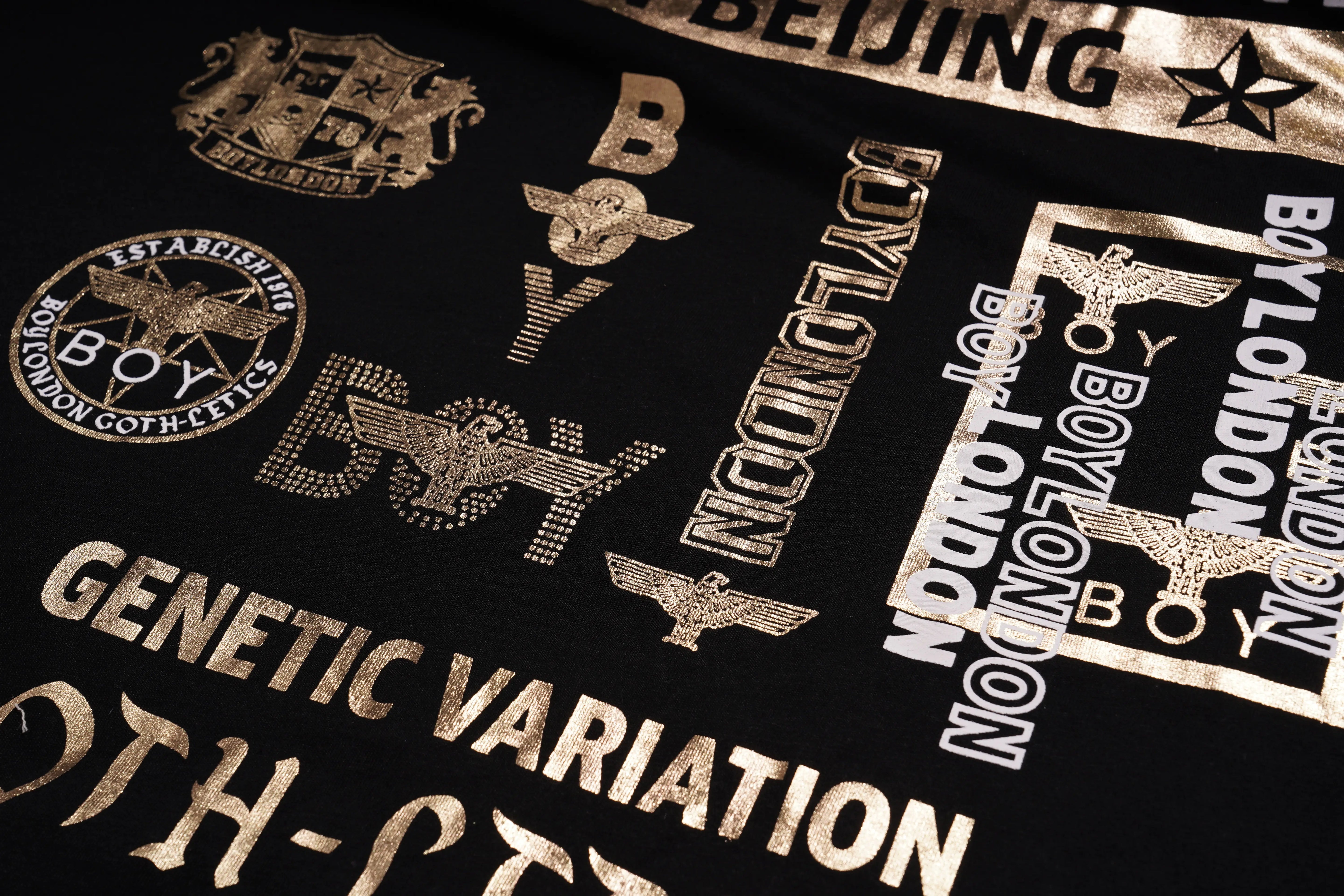 Close-up of a black fabric with gold text and logos, including 'Boy London' and 'Genetic Variation'. TCH Embroidered Floral Butterfly Rhinestone T-Shirt